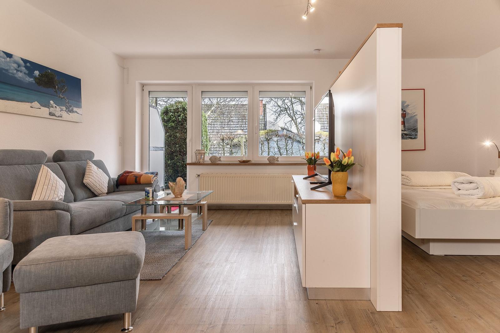 Wohnzimmer mit Sofa, Couchtisch und Fenster. Schlafzimmer mit Bett und TV-Tisch sichtbar.