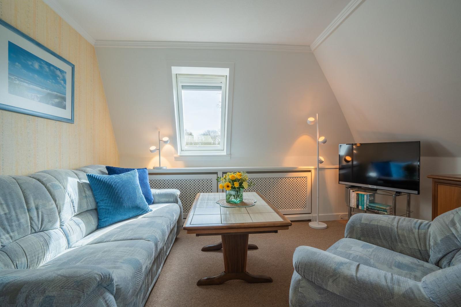 Wohnzimmer mit Sofa, Sessel, Tisch und Fernseher unter Dachschräge.
