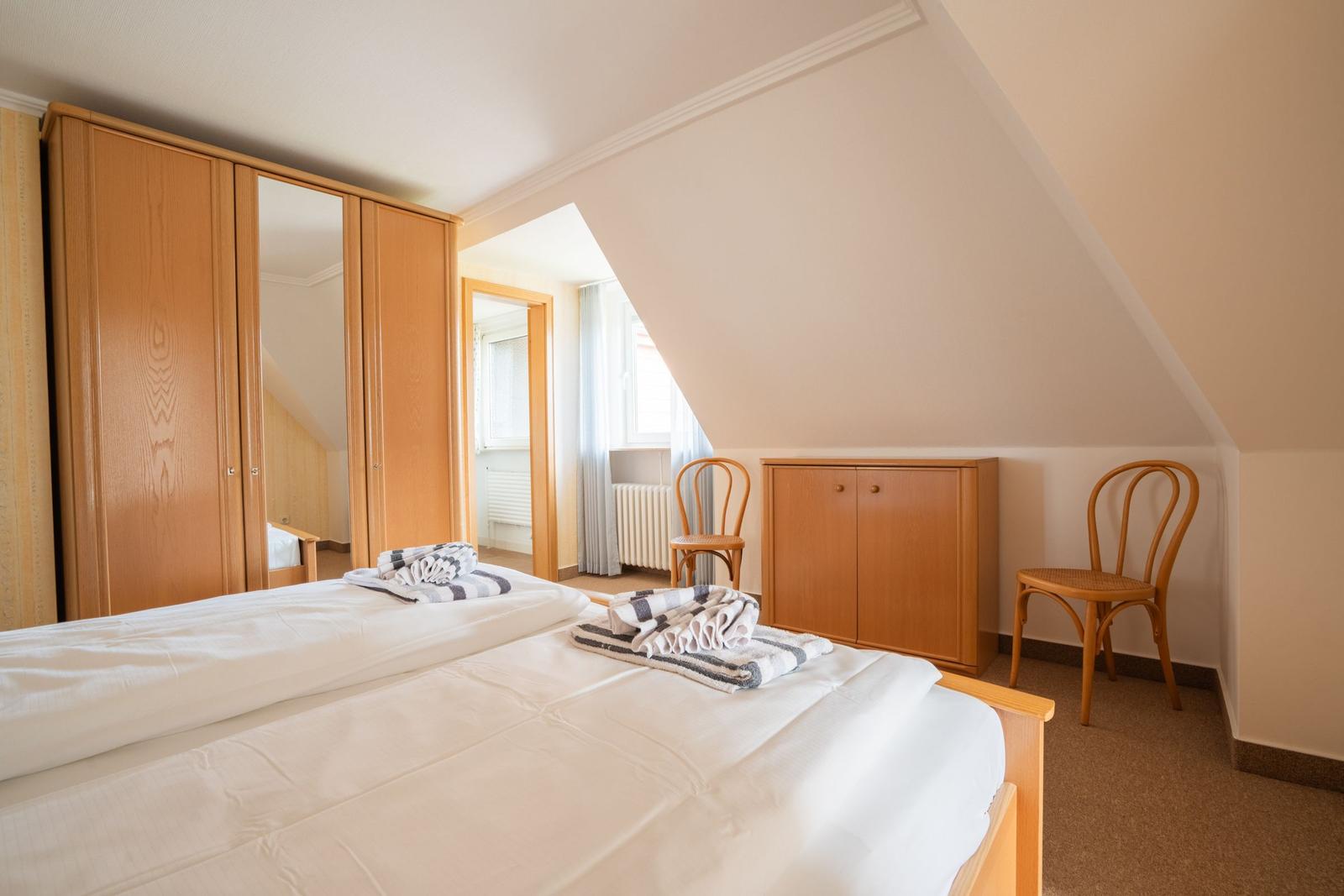 Doppelbett mit weißen Laken, Holzschrank und zwei Stühle in einem Dachzimmer.