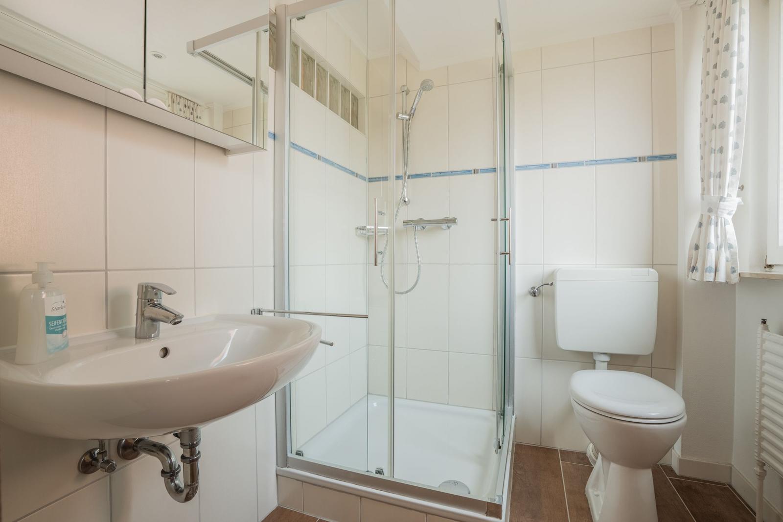 Badezimmer mit Dusche, Waschbecken und Toilette. Weiße Fliesen und brauner Boden.