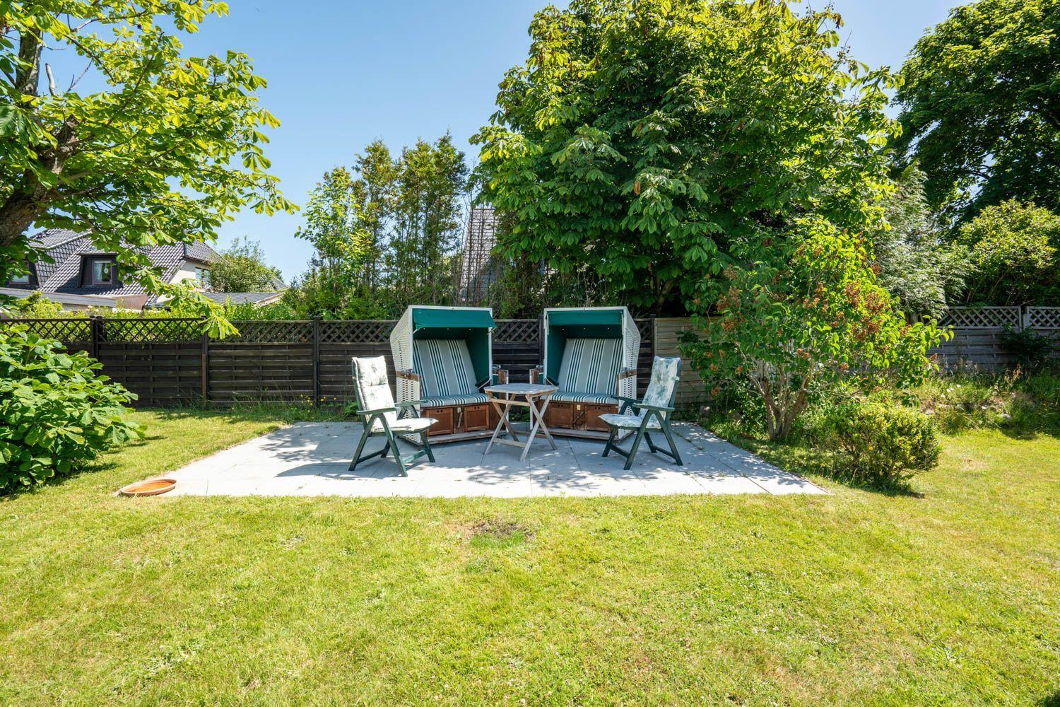Hinterhof mit zwei Strandkörben, Tisch und Stühlen auf Steinfläche. Umgeben von Gras und Bäumen.