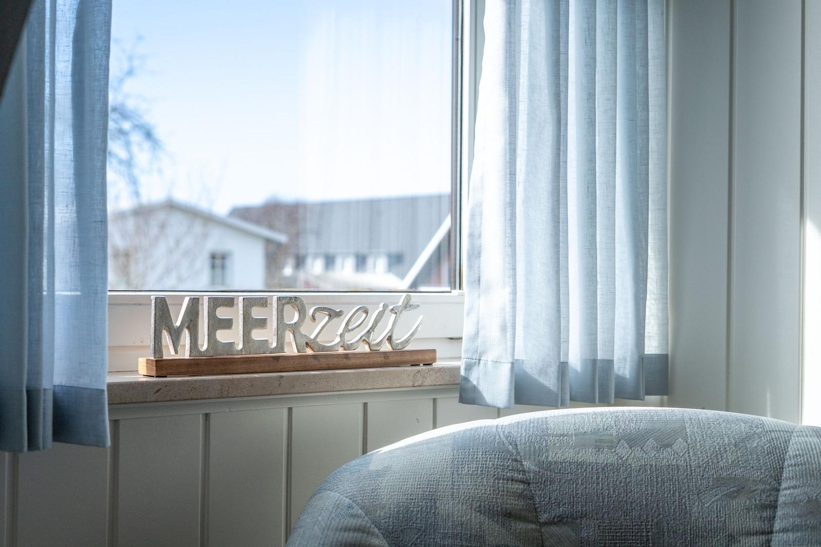 Auf dem Fensterbrett steht ein Schild mit der Aufschrift MEERZEIT. Vor dem Fenster hängen blaue Vorhänge.