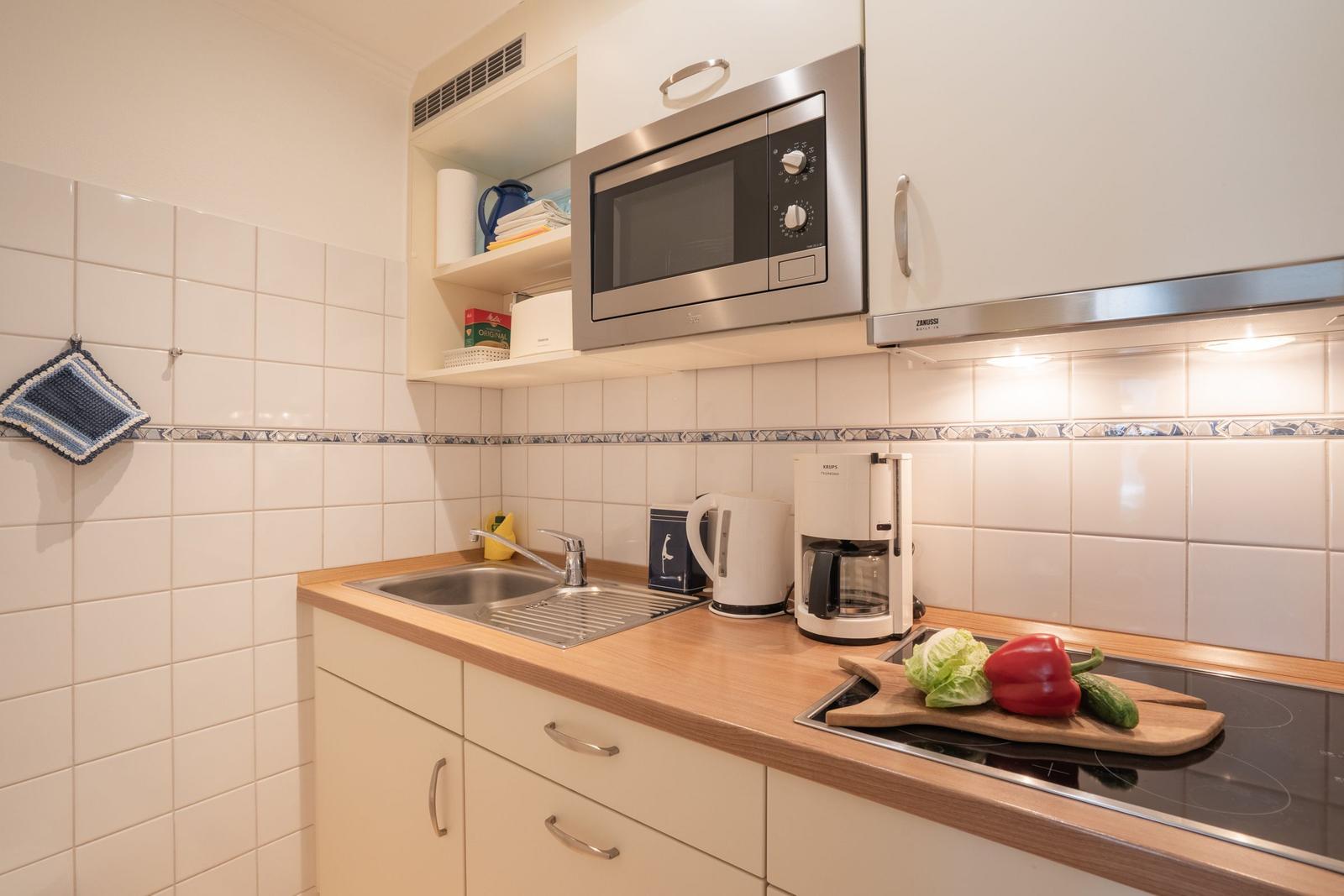 Küche mit Spüle, Mikrowelle, Kaffeemaschine und Kochfeld mit Gemüse.