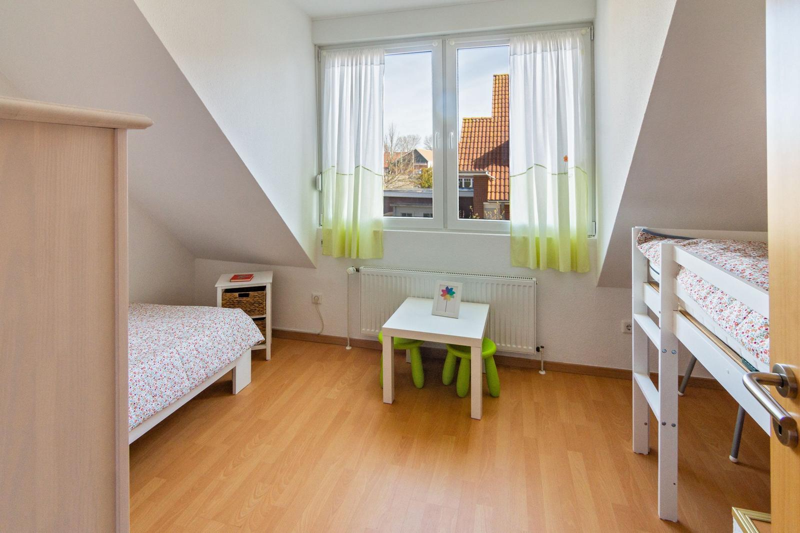 Zimmer mit Dachschrägen, Einzelbett, Doppelstockbett, Tisch und Stühlen, Fenster mit Vorhängen.