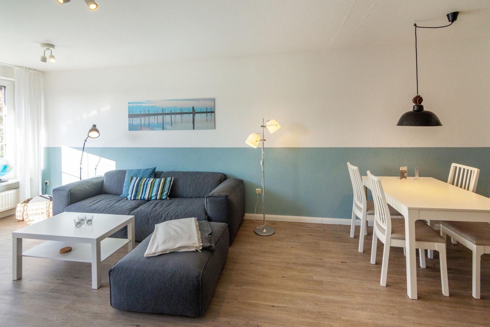 Wohn- und Essbereich mit grauem Sofa, weißem Tisch und Stühlen, blau-weißen Wänden und Holzboden.