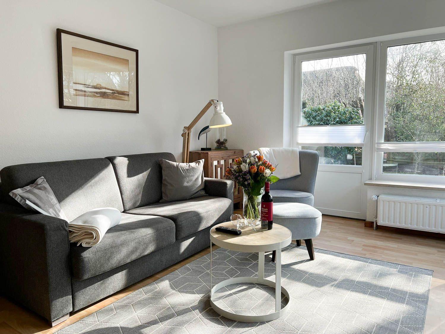 Wohnzimmer mit Sofa, Sessel und Tisch. Fensterblick auf Garten. Wandbild und Lampe.