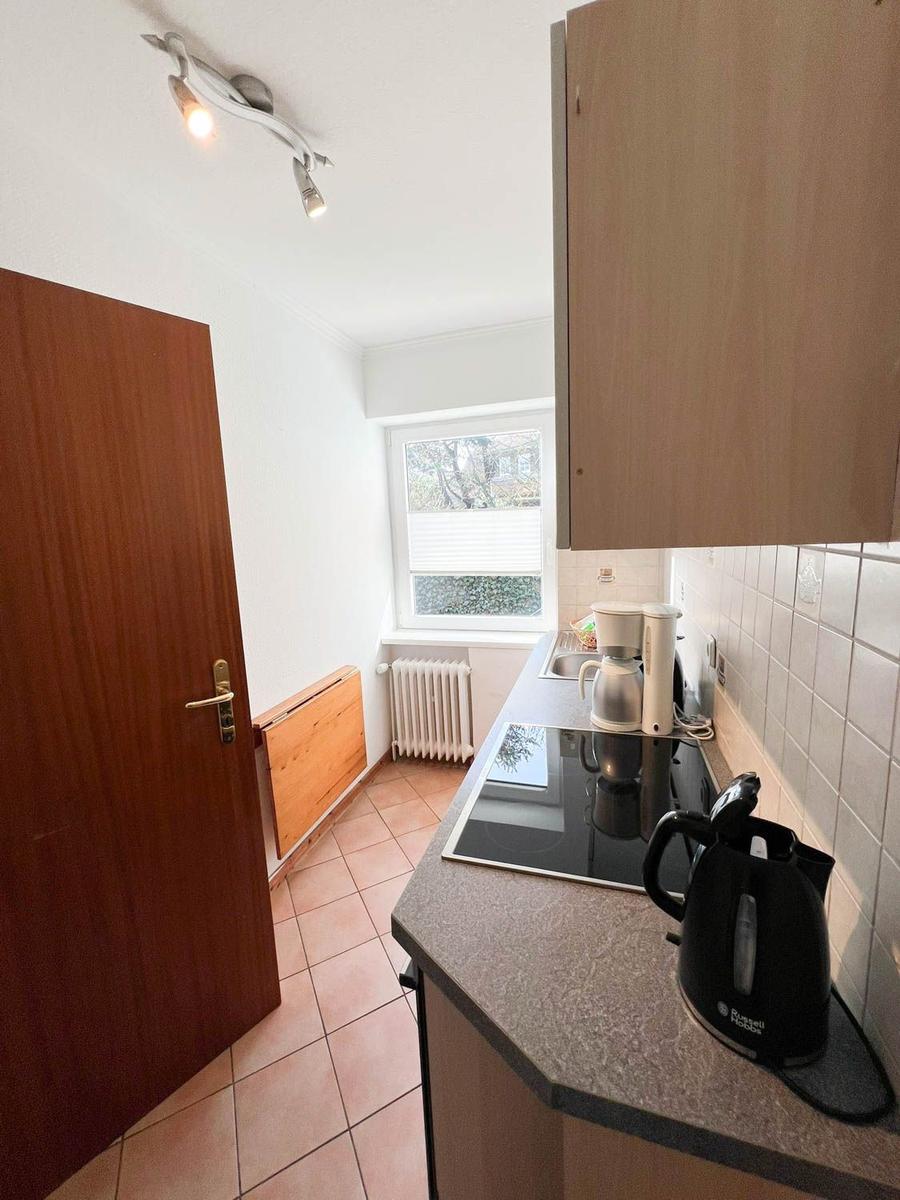 Küche mit Arbeitsplatte, Herd, Kaffeemaschine und Kanne. Fenster mit Jalousie und Heizkörper.