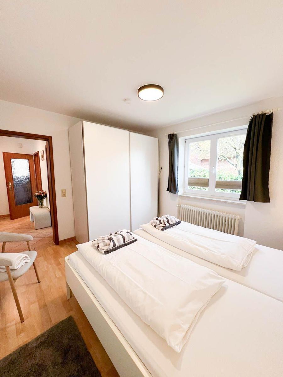 Zimmer mit Doppelbett, Schrank und Fenster. Holzboden und Stuhl sind sichtbar.