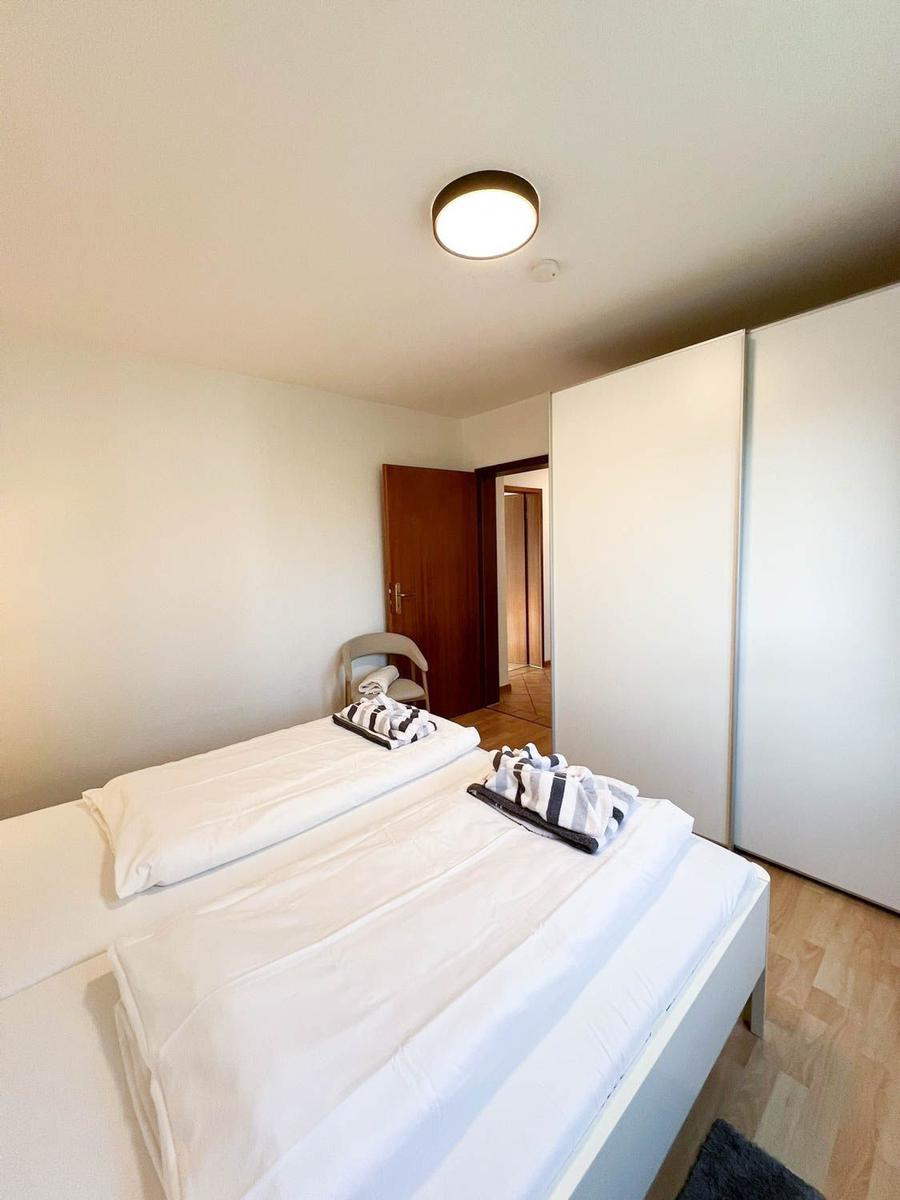 Schlafzimmer mit Bett, Kleiderschrank und Stuhl. Boden aus Holz, Decke mit Lampe.