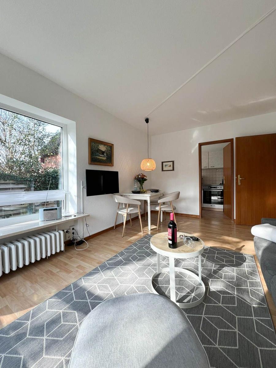 Wohn- und Essbereich mit Küche im Hintergrund. Fenster, TV, Tisch mit Stühlen und Couch.