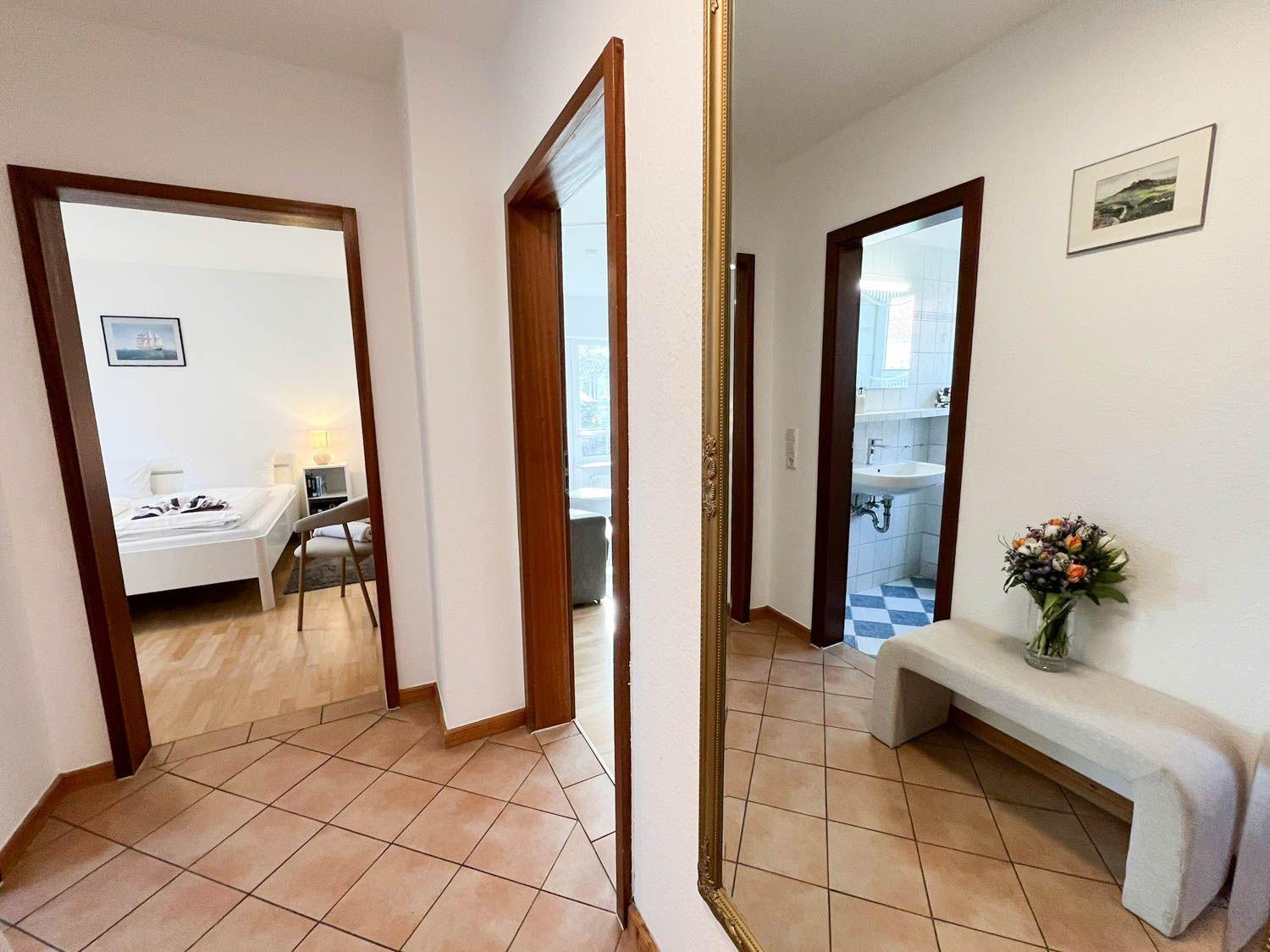 Flur mit Blick in Schlafzimmer, Bad und Wohnbereich. Holzrahmen, Fliesenboden und Blumen.