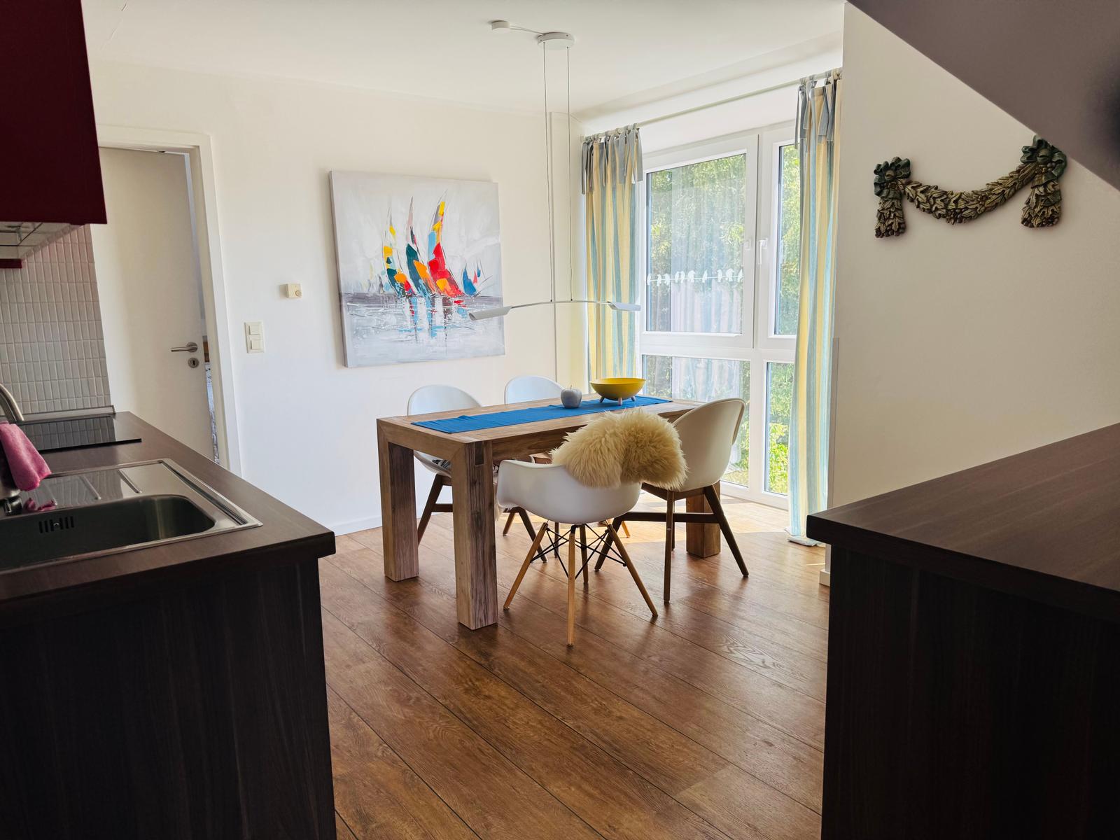 Essbereich mit Holztisch, weißen Stühlen und großem Fenster. Küche mit Arbeitsplatte und Spüle.