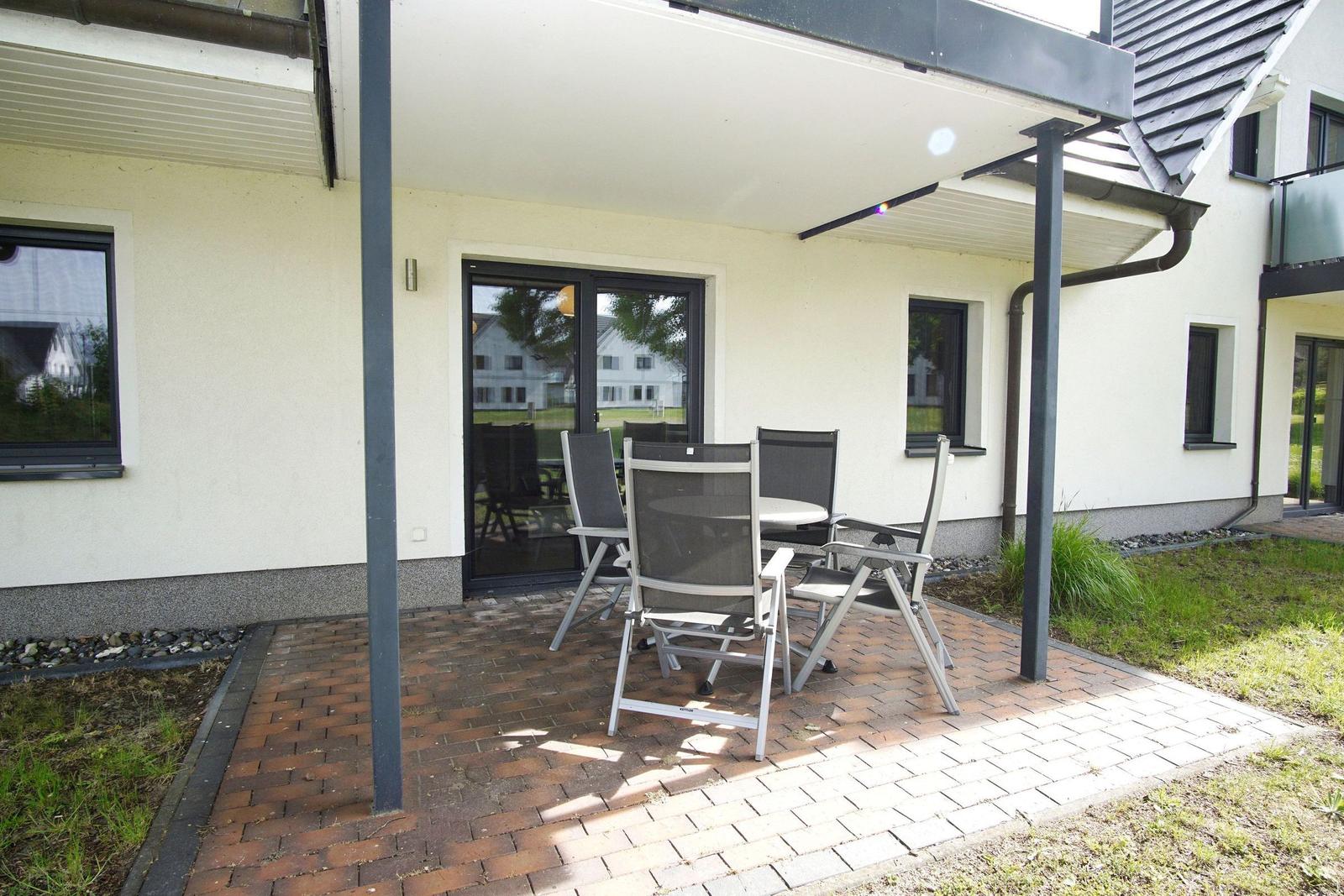 Terrasse mit Sitzgruppe unter Überdachung. Holz- und Steinbelag. Fenster und Tür sichtbar.
