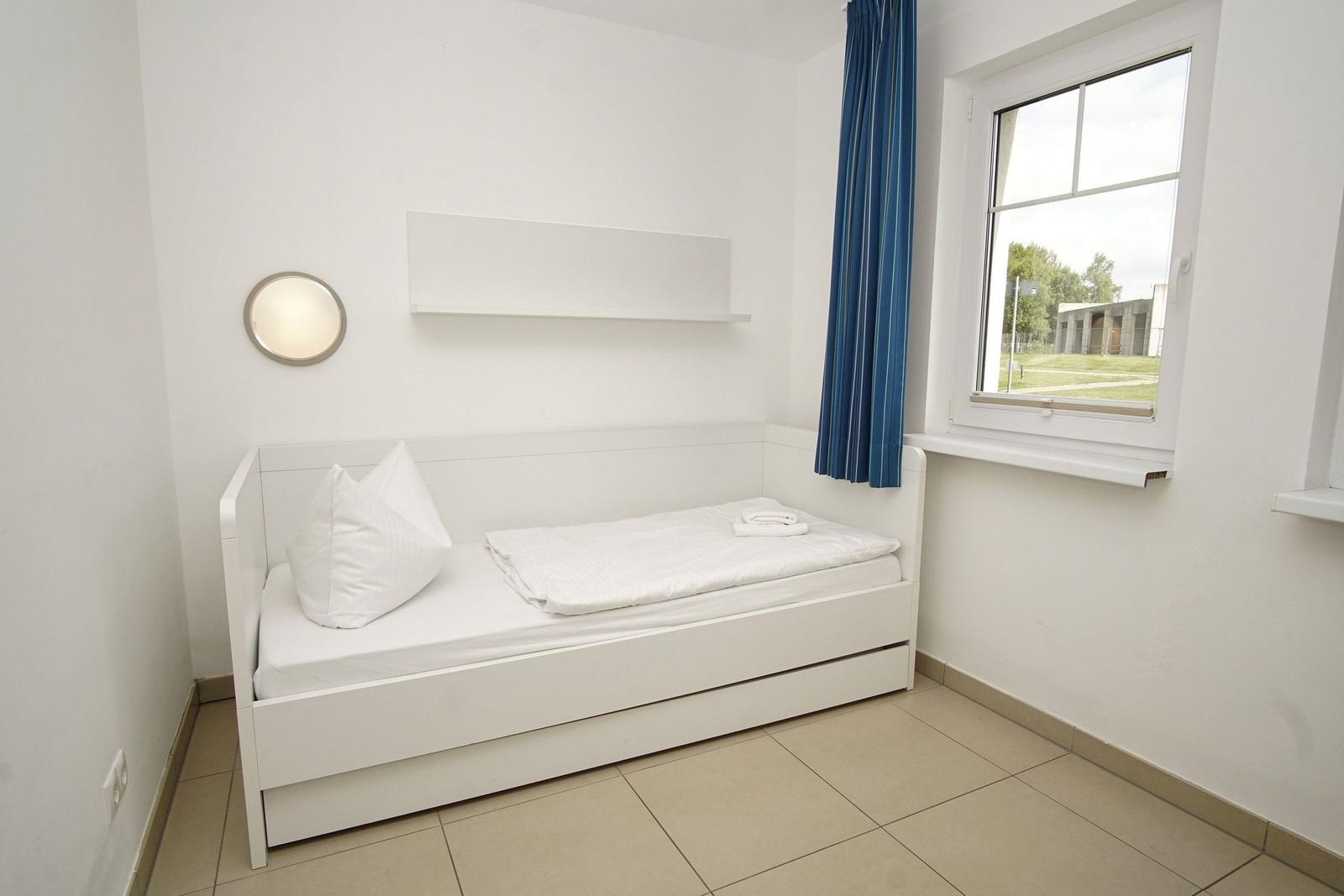 Ein Schlafzimmer mit weißem Bett, blauen Vorhängen und Fensterblick auf Grünfläche.