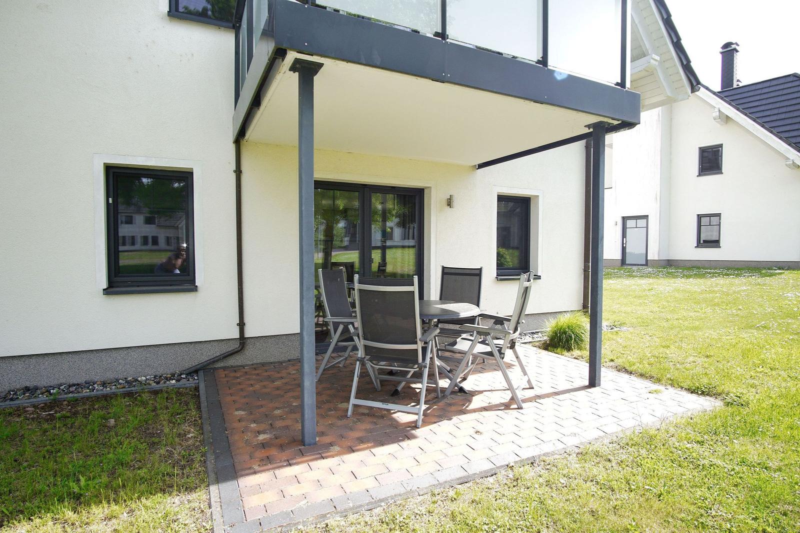 Terrasse mit Tisch und Stühlen unter Überdachung. Hintergrund: weißes Haus mit Grünfläche.