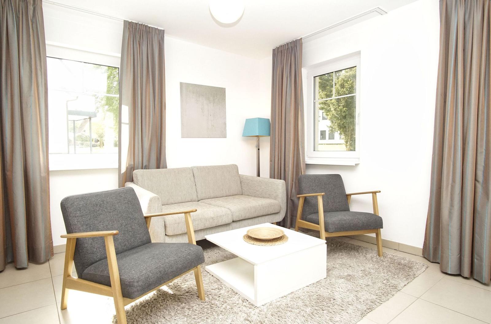 Wohnzimmer mit Sofa, zwei Stühlen und Tisch. Fenster mit Vorhängen. Wandbild und Stehlampe.