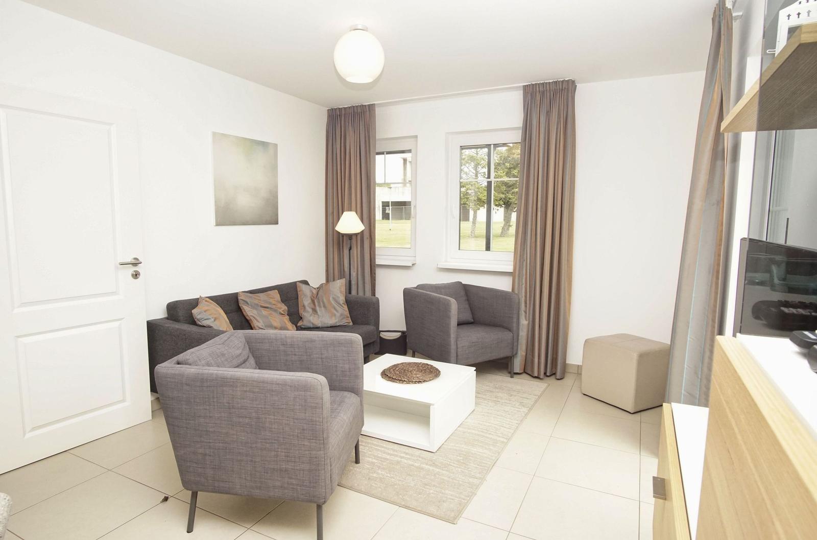 Living room with sofa, armchairs, and windows overlooking a green area.