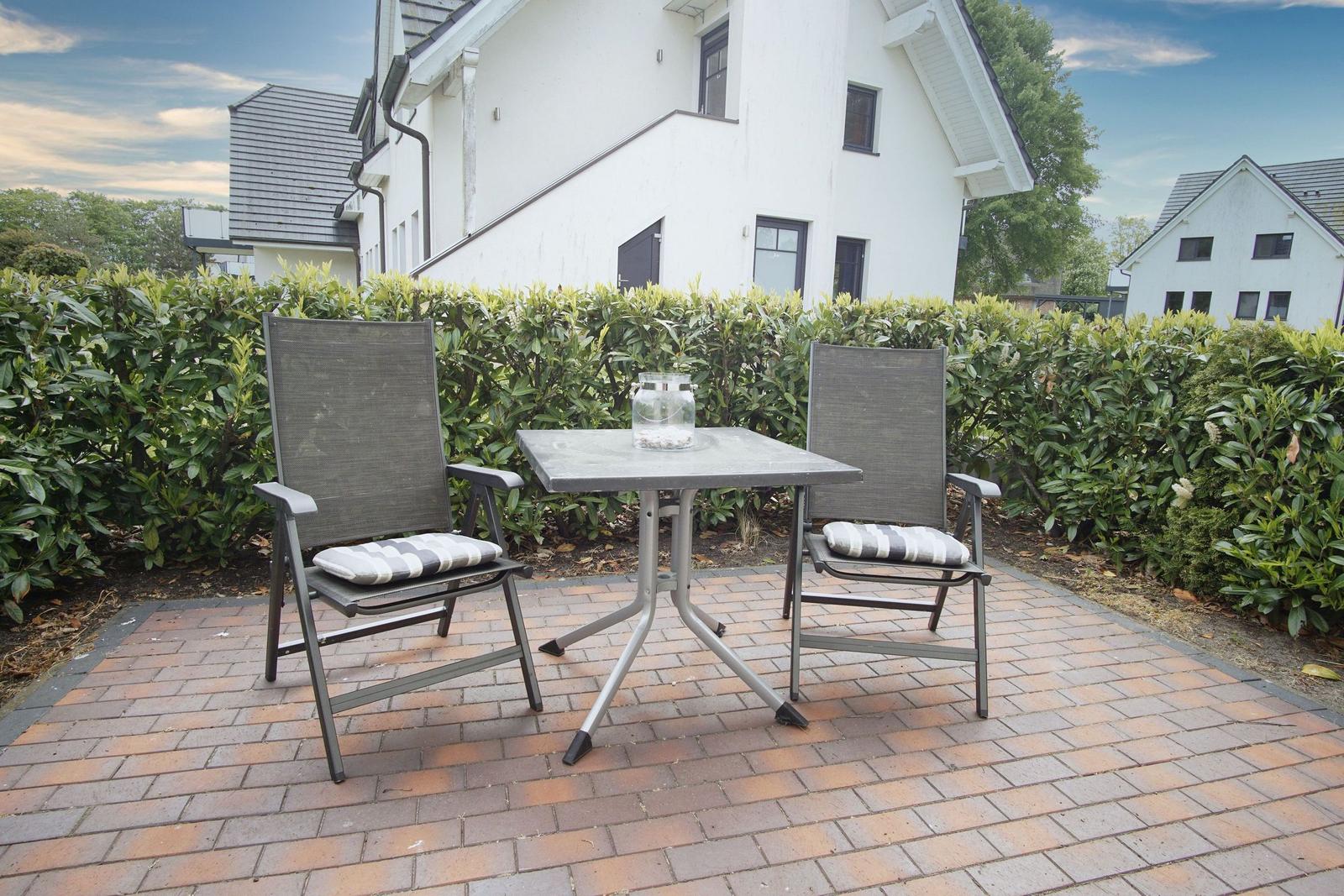 Terrace with table and two chairs in front of white house with hedge.