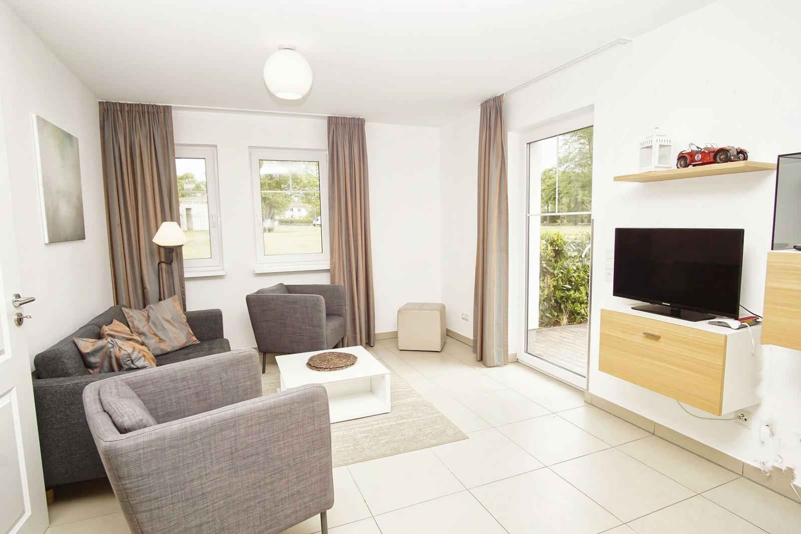 Living room with sofa, armchairs, coffee table, and TV. Door and windows with curtains.