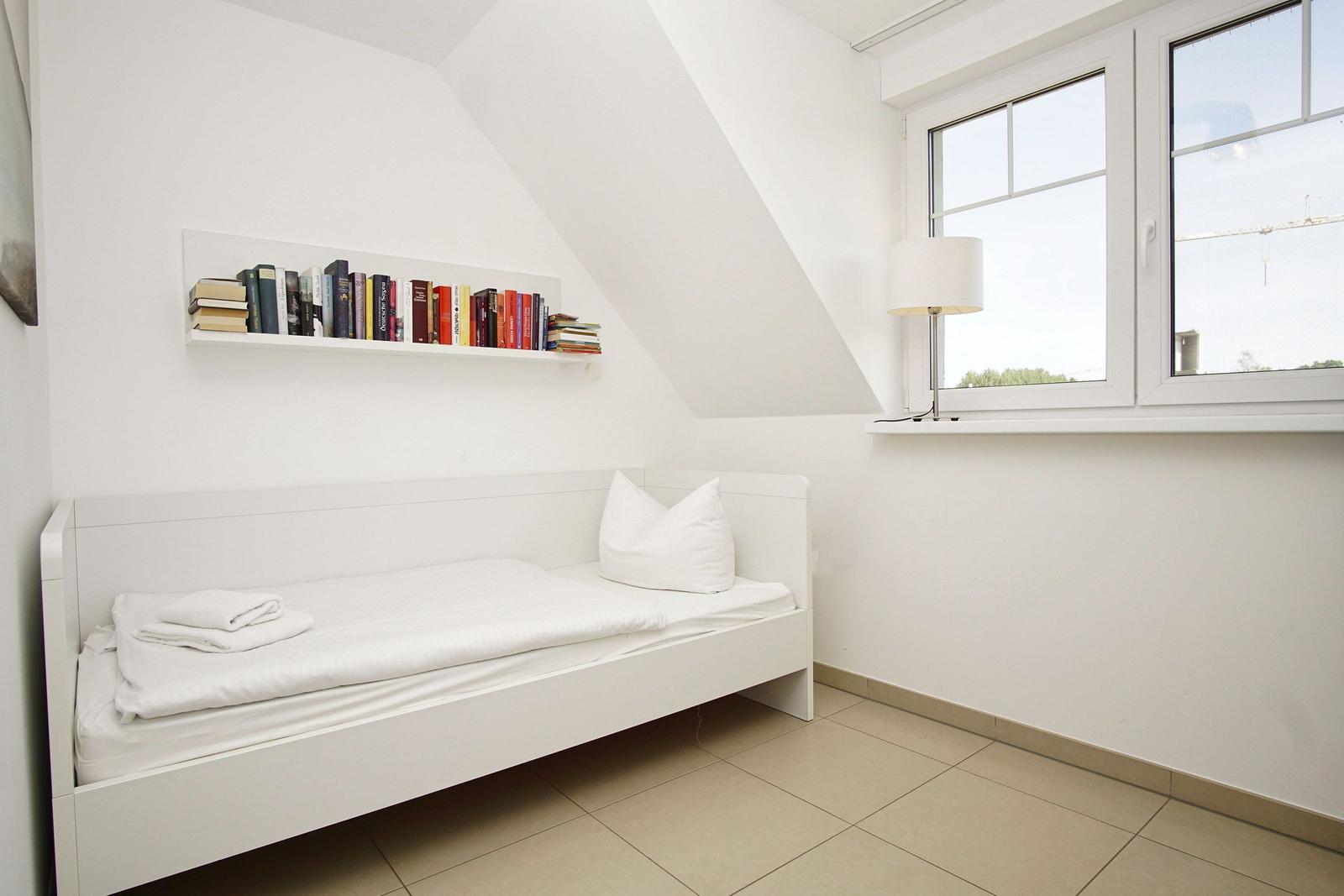 Schlafzimmer mit weißem Bett, Bücherregal und Fenster.