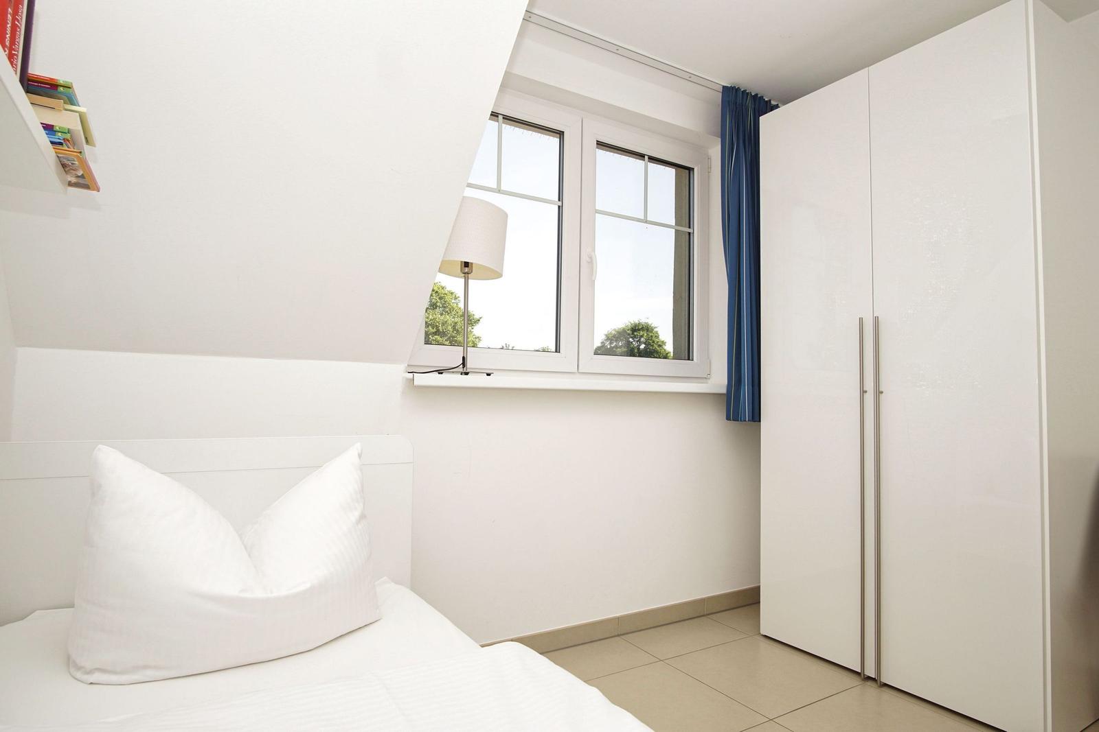 Schlafzimmer mit weißem Bett, Fenster und Kleiderschrank.
