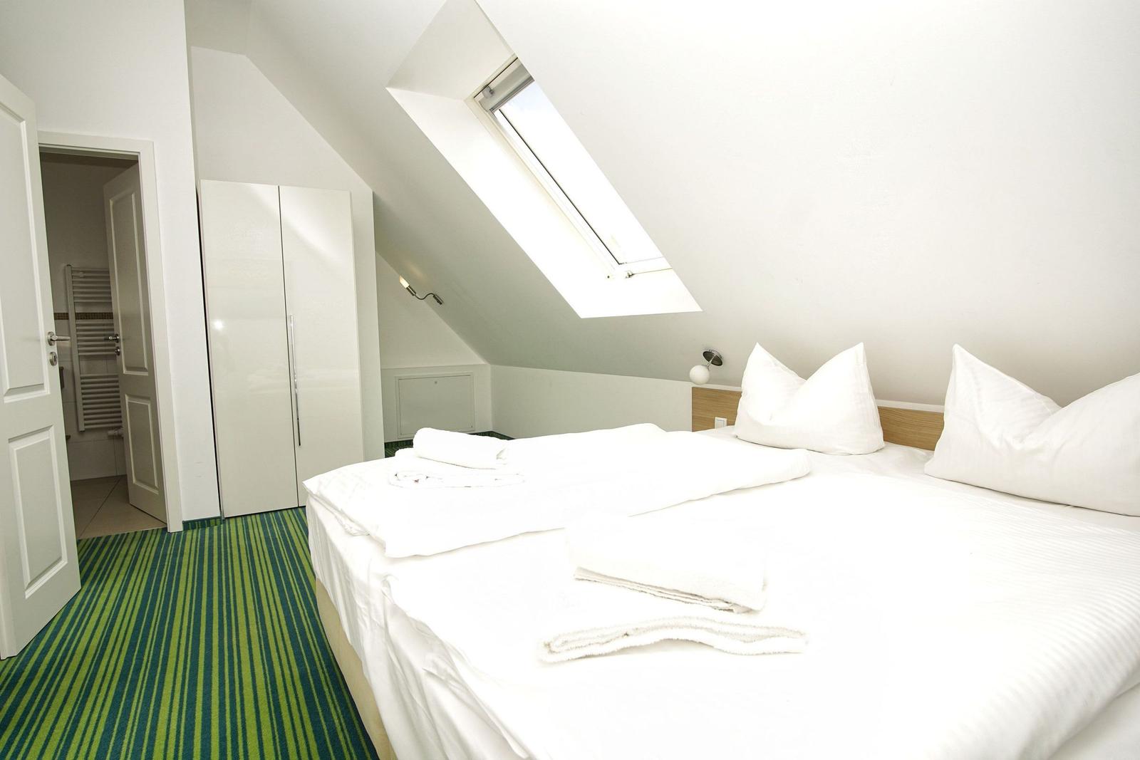 Schlafzimmer mit Dachfenster, Bett und Kleiderschrank. Grün-weißer Teppich. Tür zum Bad.
