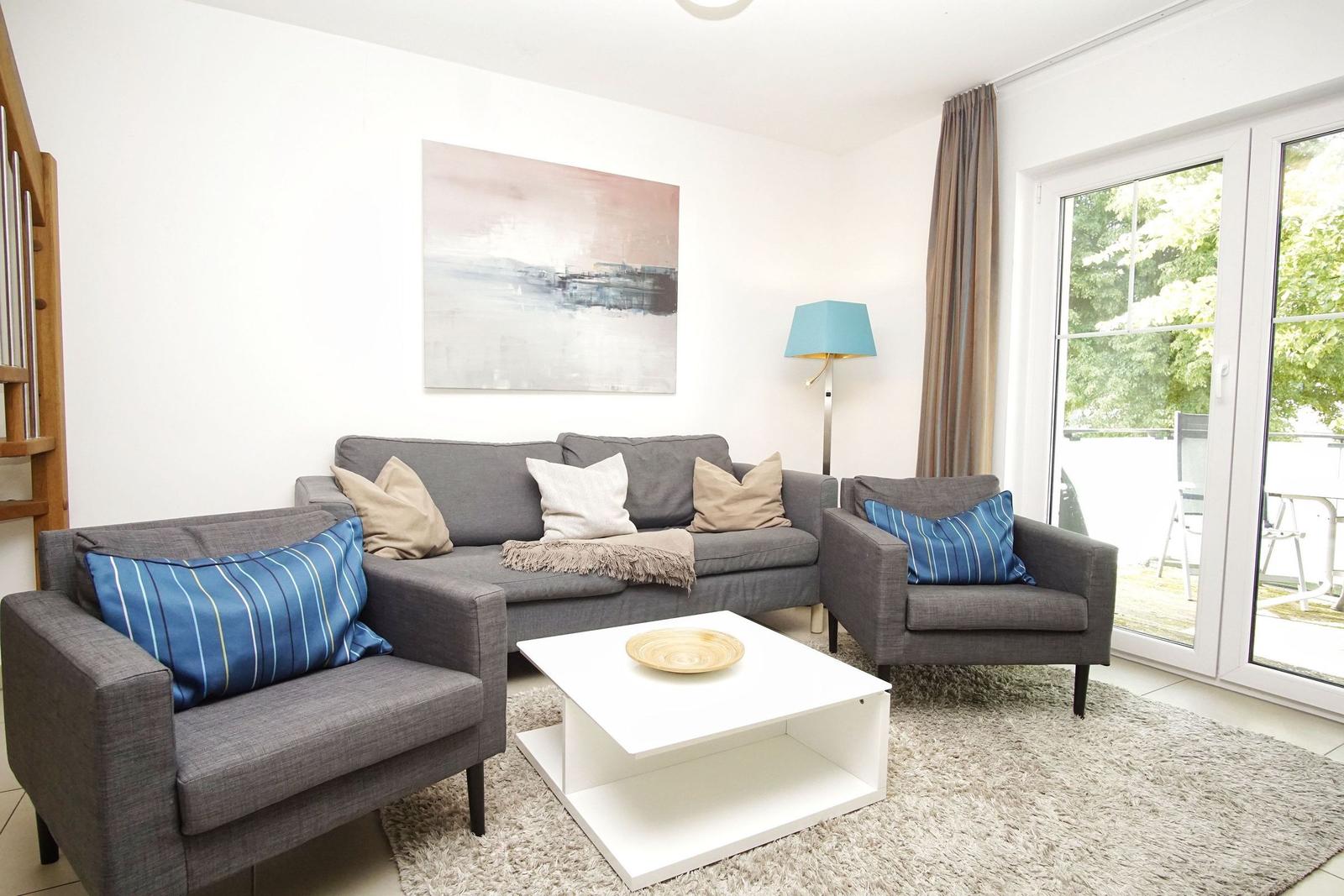 Wohnzimmer mit grauem Sofa, zwei Sesseln und weißem Couchtisch. Große Glastüren führen auf die Terrasse.