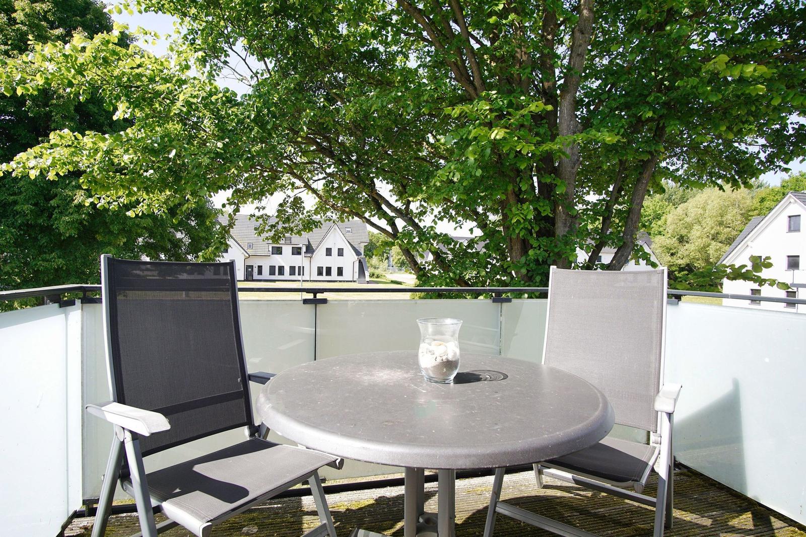 Terrasse mit Tisch und zwei Stühlen, umgeben von Bäumen und Häusern im Hintergrund.