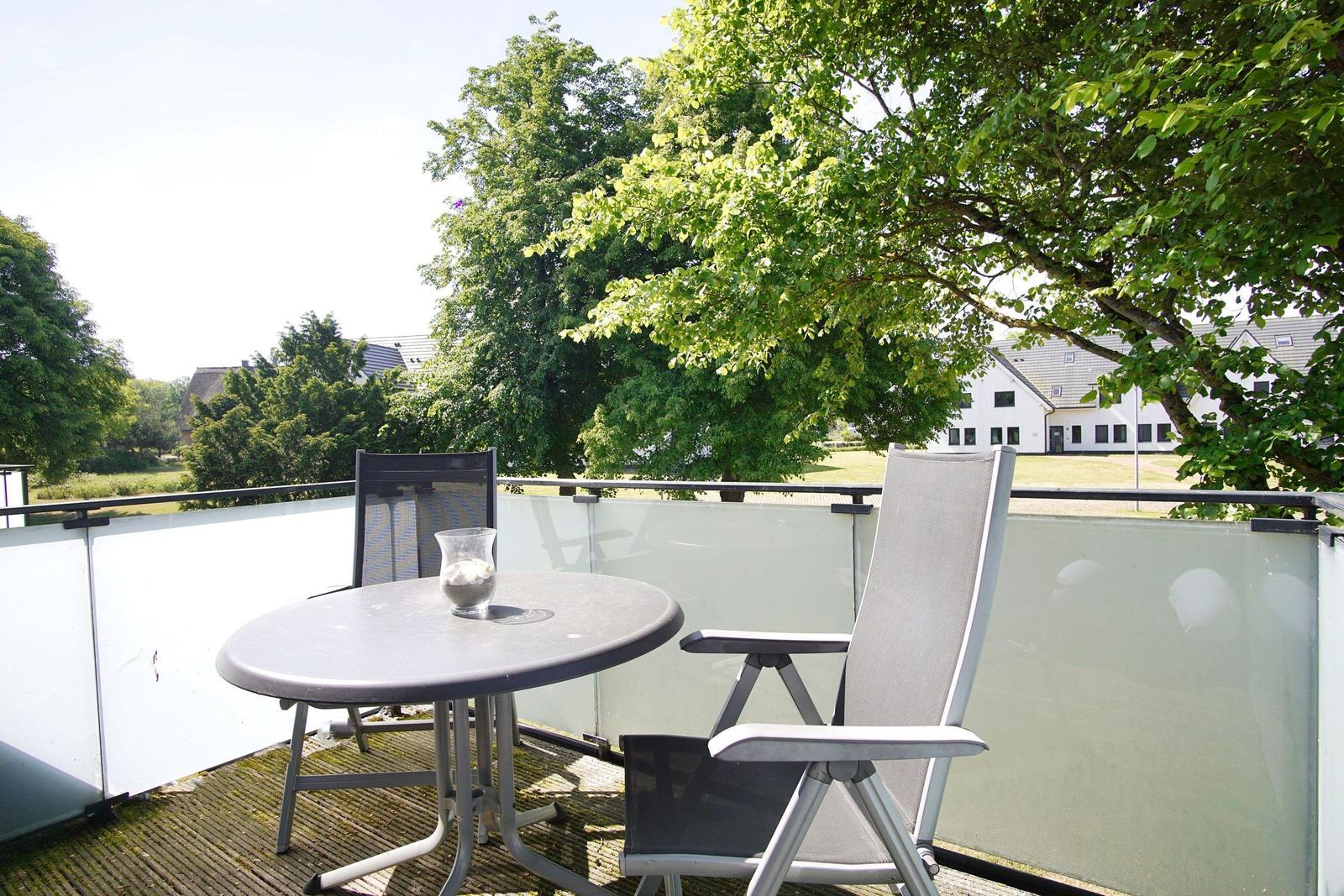 Terrasse mit Tisch und zwei Stühlen. Blick auf grüne Bäume und ein Haus.