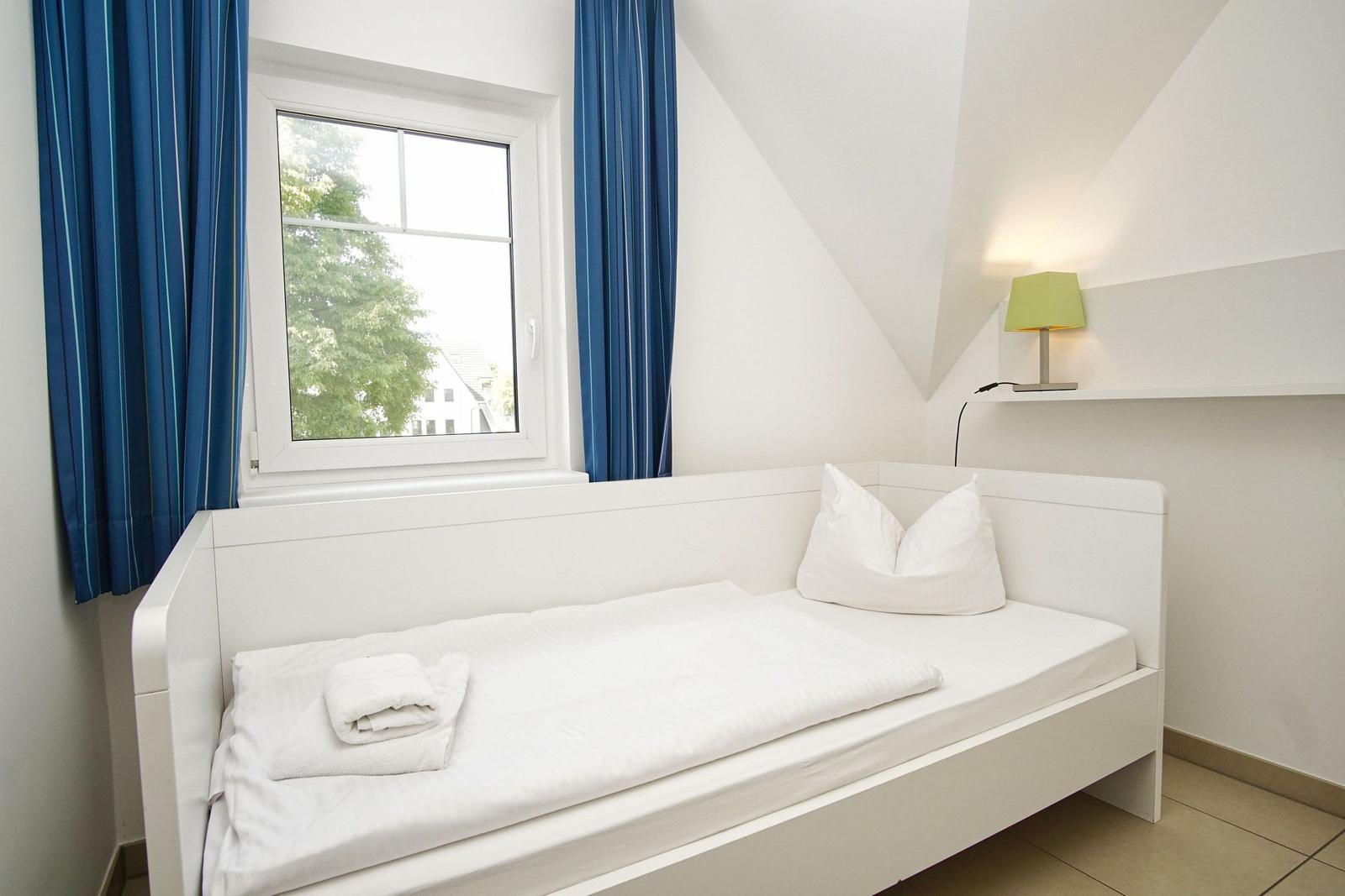 Schlafzimmer mit weißem Bett, blauen Vorhängen und Fensterblick auf Grün.