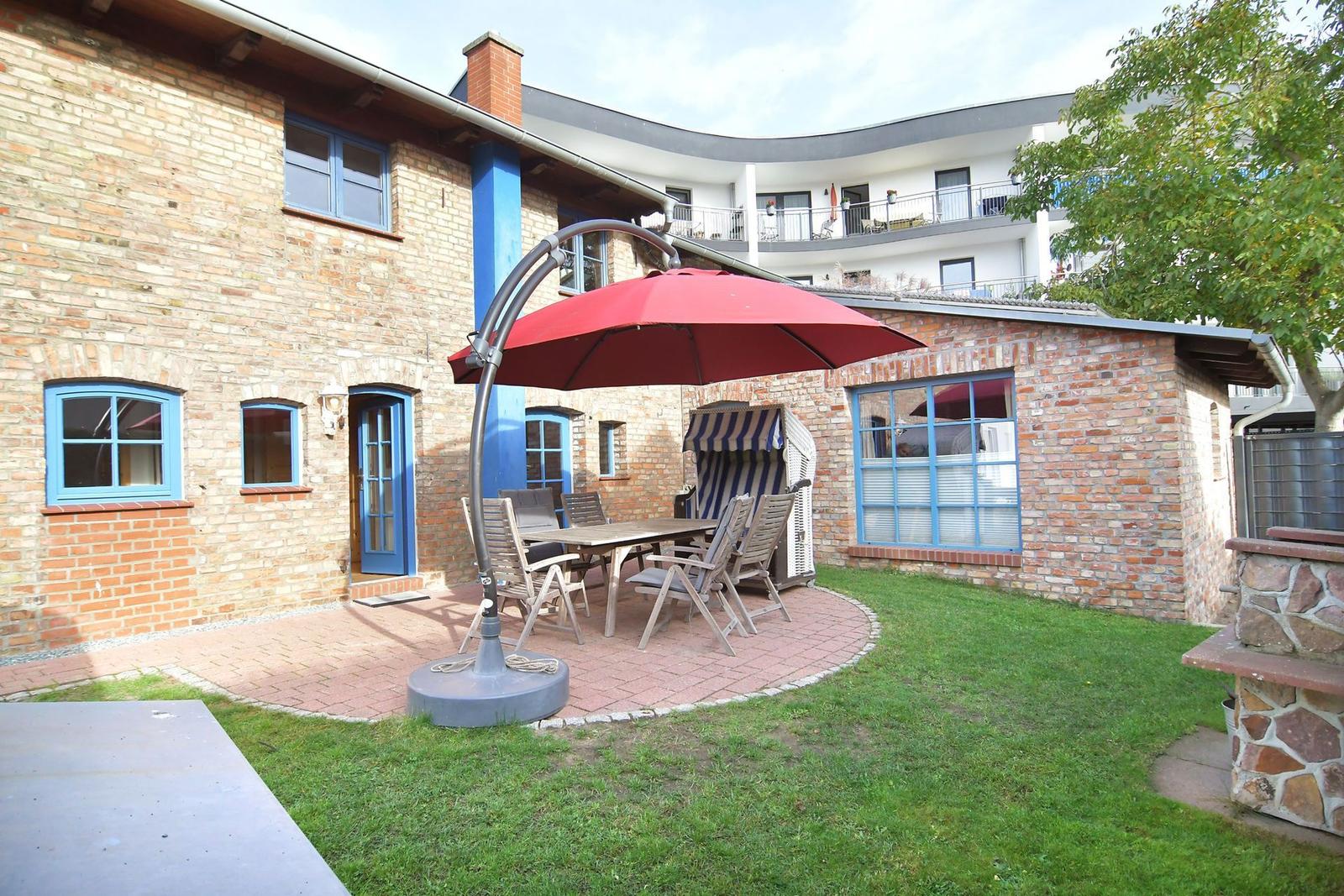 Hinterhof mit Terrasse, Tisch, Stühlen und rotem Sonnenschirm. Backsteinhaus mit blauen Fenstern.