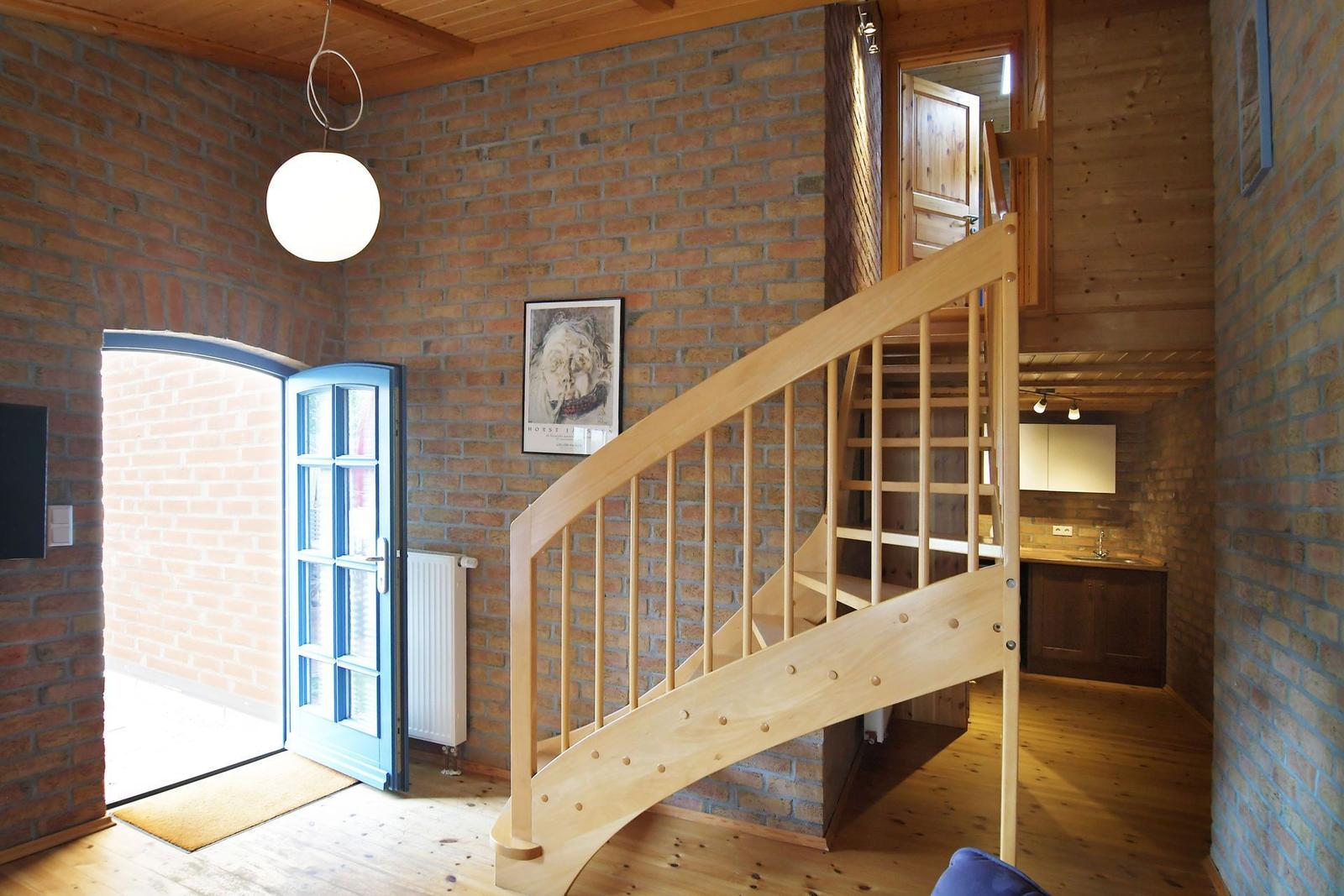 Holzgetreppe führt zur oberen Etage. Ziegelwand, blaue Tür und Küchenbereich sichtbar.