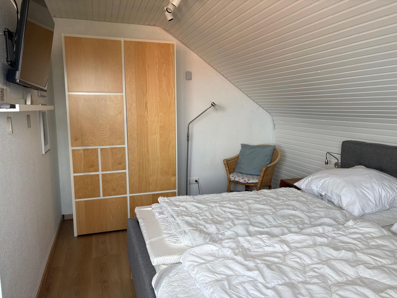 Schlafzimmer mit Bett, Kleiderschrank und Sessel unter schrägem Dach.