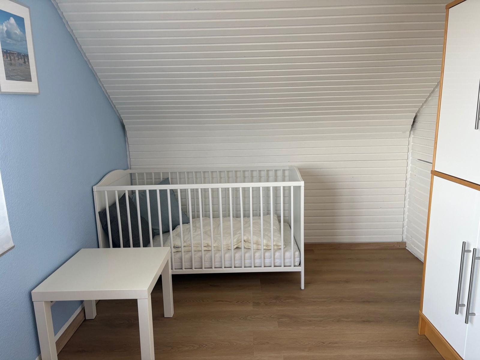 Kinderzimmer mit weißem Babybett, Tisch und Schrank. Dachsatteldach und blaue Wand.