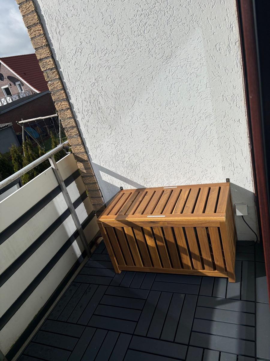 Holzbox auf Balkon mit dunkler Terrassenplatte und weißer Wand.