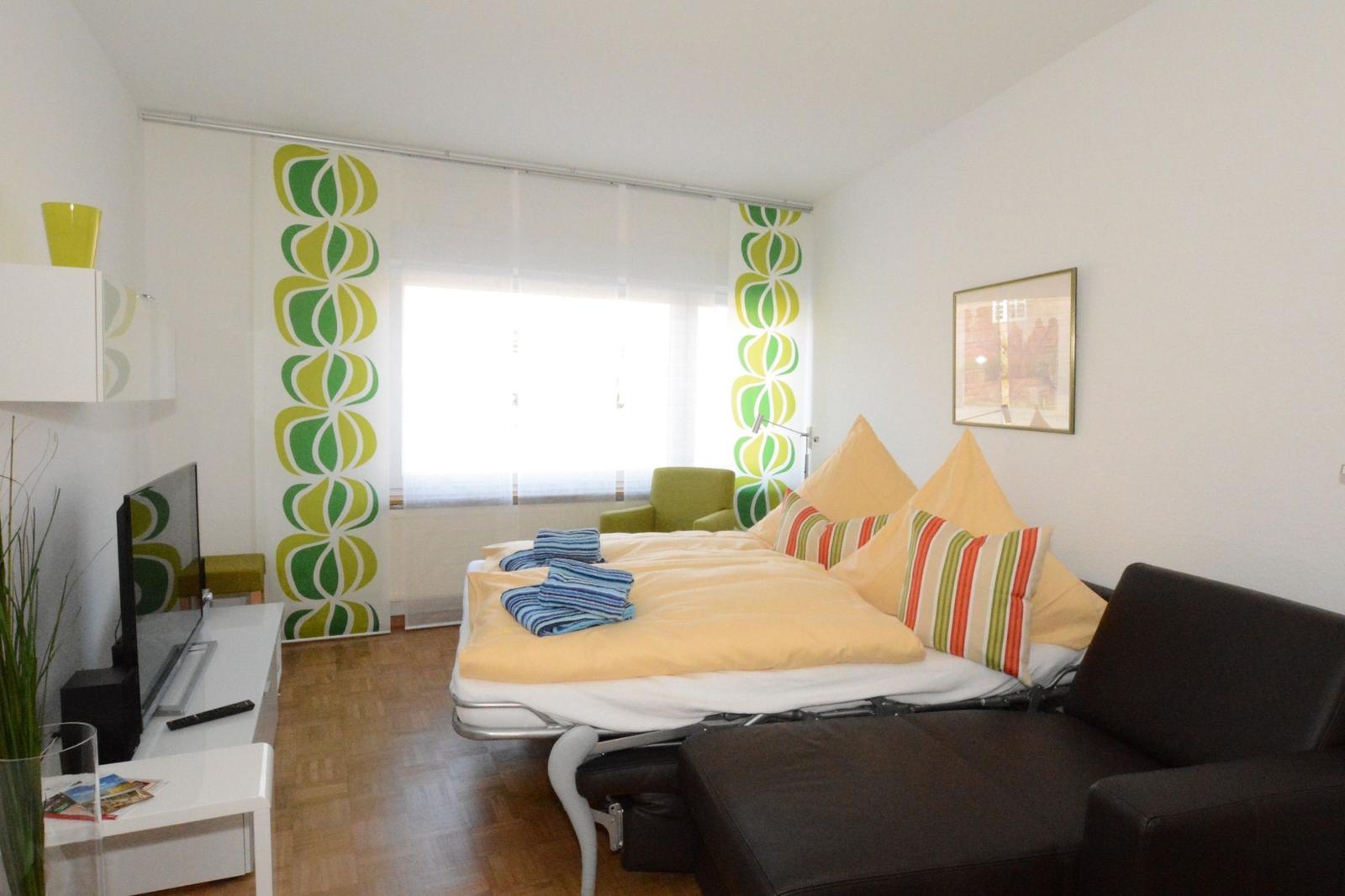 Zimmer mit Bett, Sofa, TV und Fenster mit grün-gelben Vorhängen.