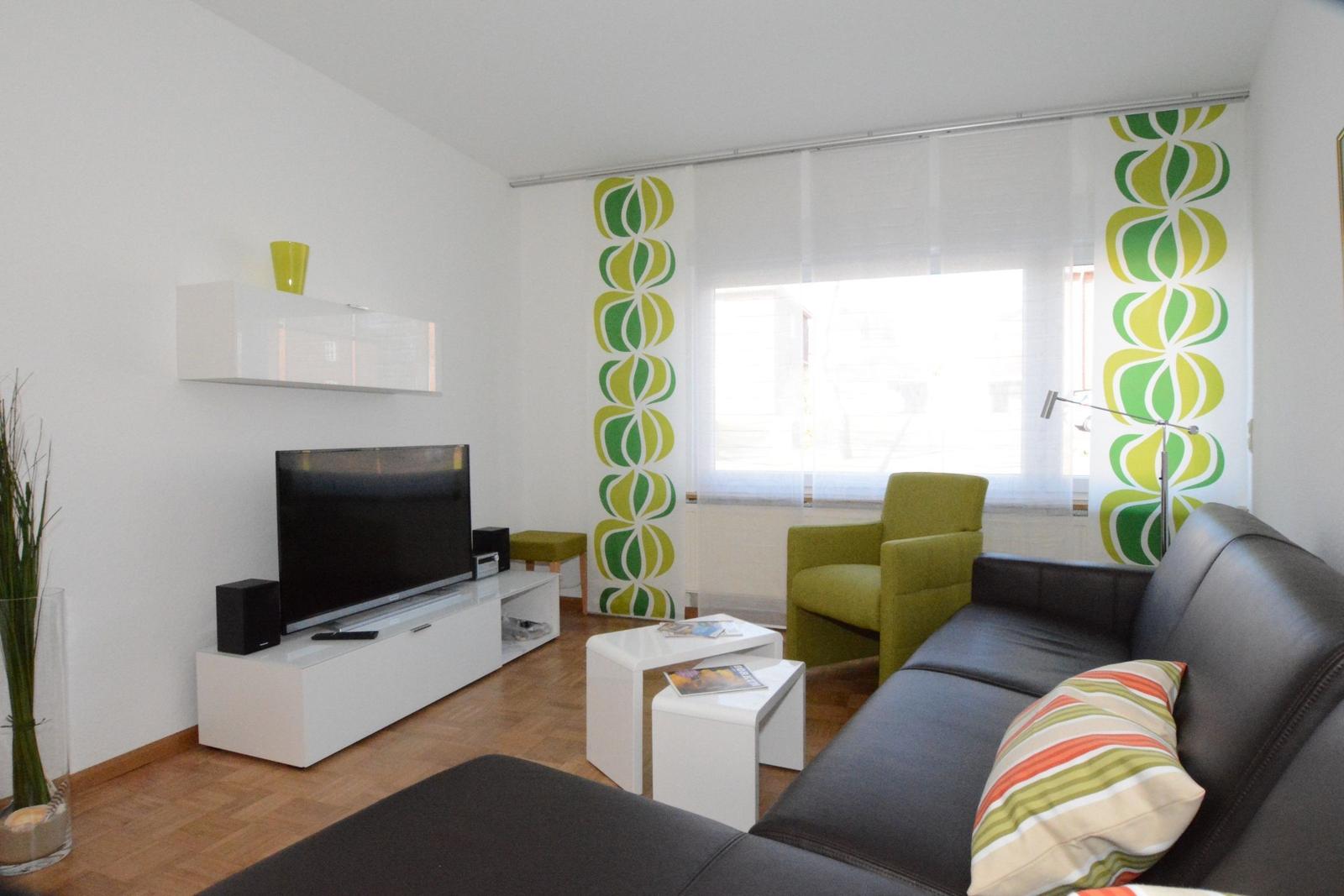 Wohnzimmer mit Sofa, Sessel, Fernseher und dekorativen Gardinen.