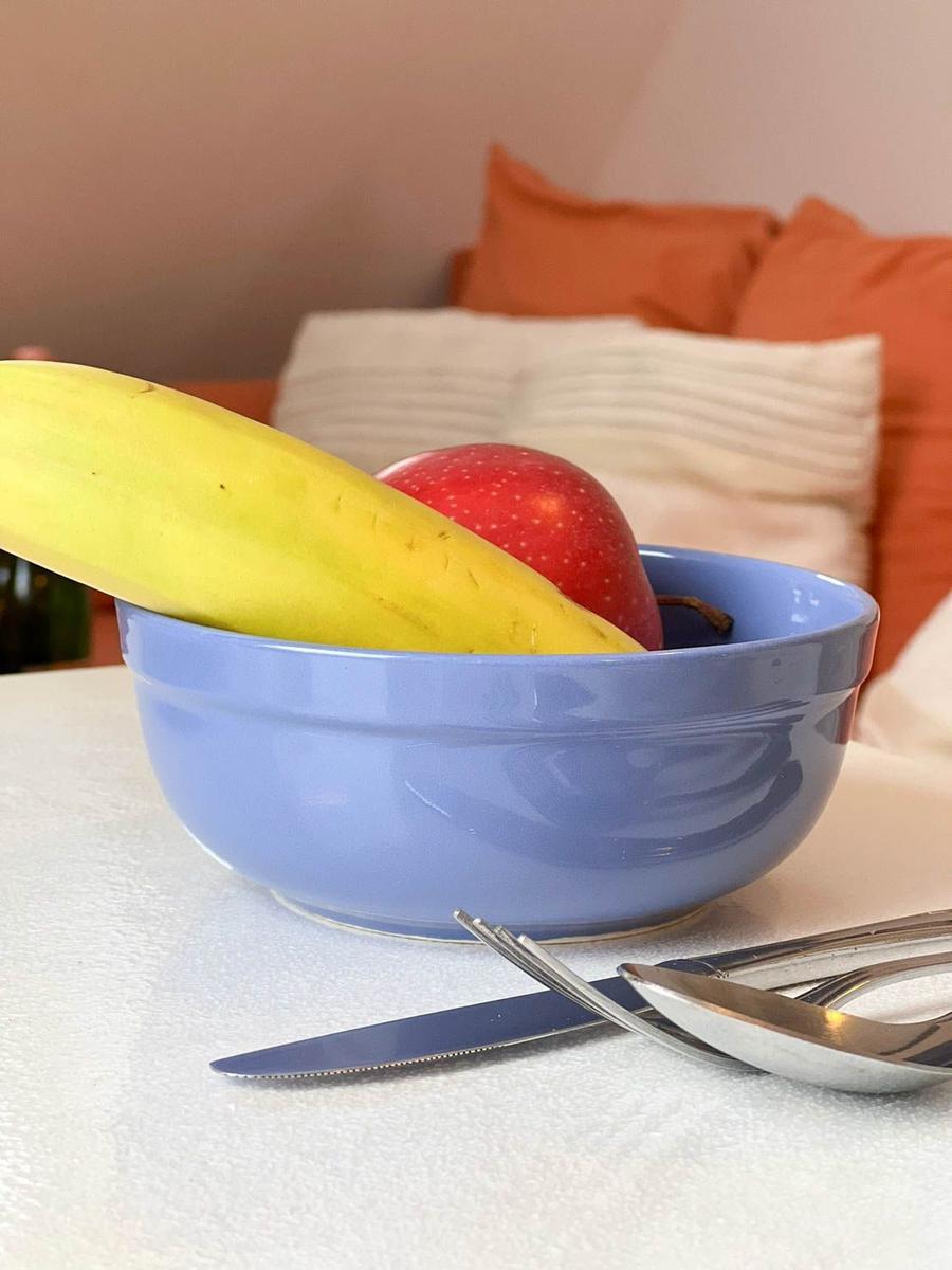 Eine blaue Schale mit Banane und Apfel auf Tisch vor Bett mit Kissen.