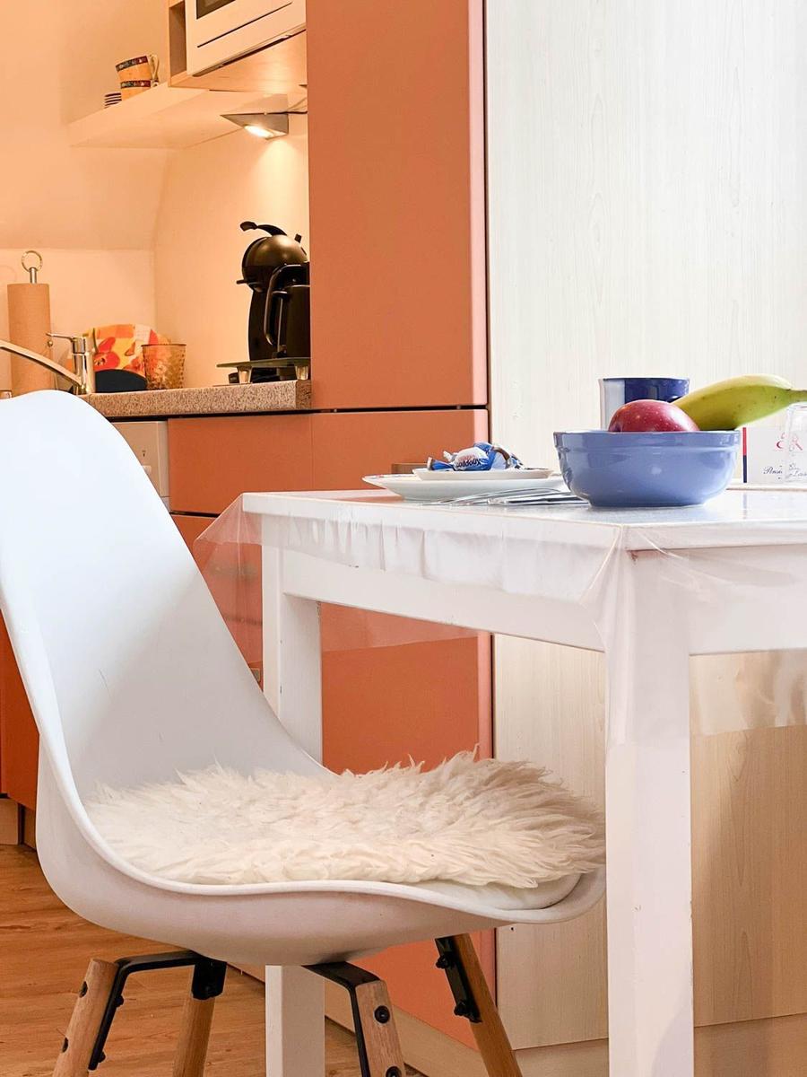 Weißer Stuhl mit Fellkissen am Esstisch. Küchenbereich mit orangefarbenen Schränken.