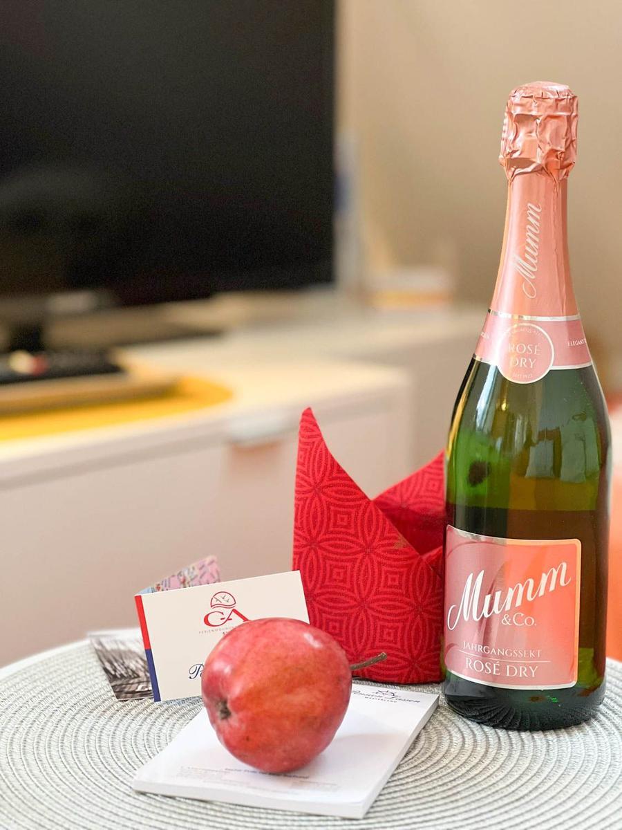 Auf einem Tisch steht eine Flasche Sekt, eine rote Frucht und ein roter Stoffbeutel.