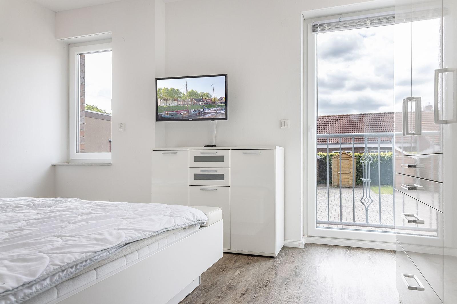 Schlafzimmer mit Bett, TV und Balkonzugang. Weiße Einrichtung und Holzboden.