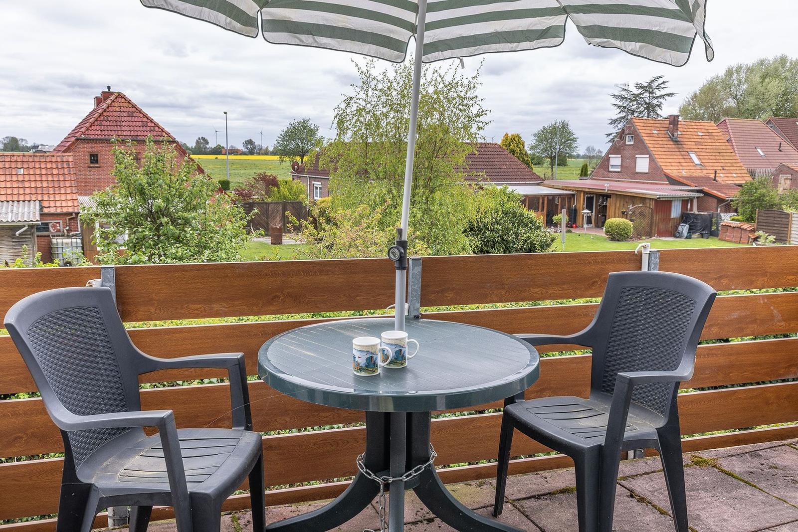 Terrasse mit Tisch, zwei Stühlen und Sonnenschirm. Blick auf Wohnhäuser und Grünflächen.