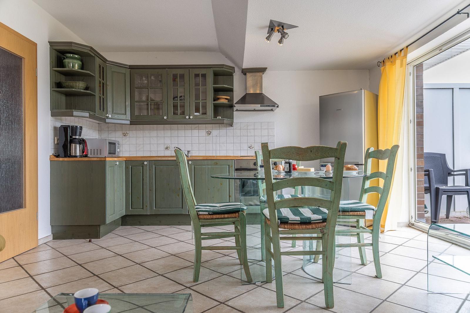 Küche mit grünen Schränken, Glas-Tisch und Stühlen. Durchgang zur Terrasse mit gelben Vorhängen.