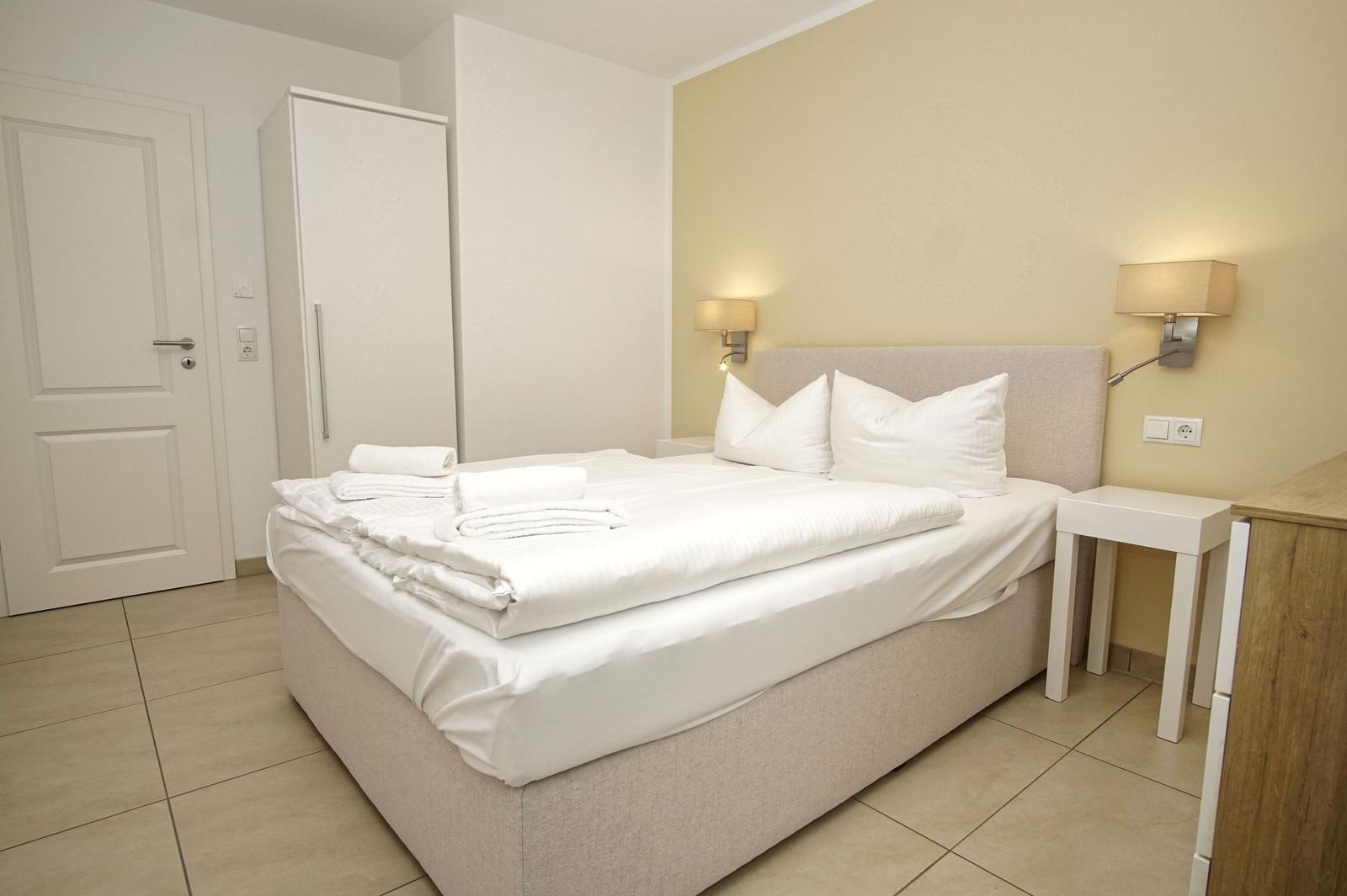 Schlafzimmer mit Bett, Nachttisch und Schrank. Wandlampen und gefaltete Handtücher sind sichtbar.
