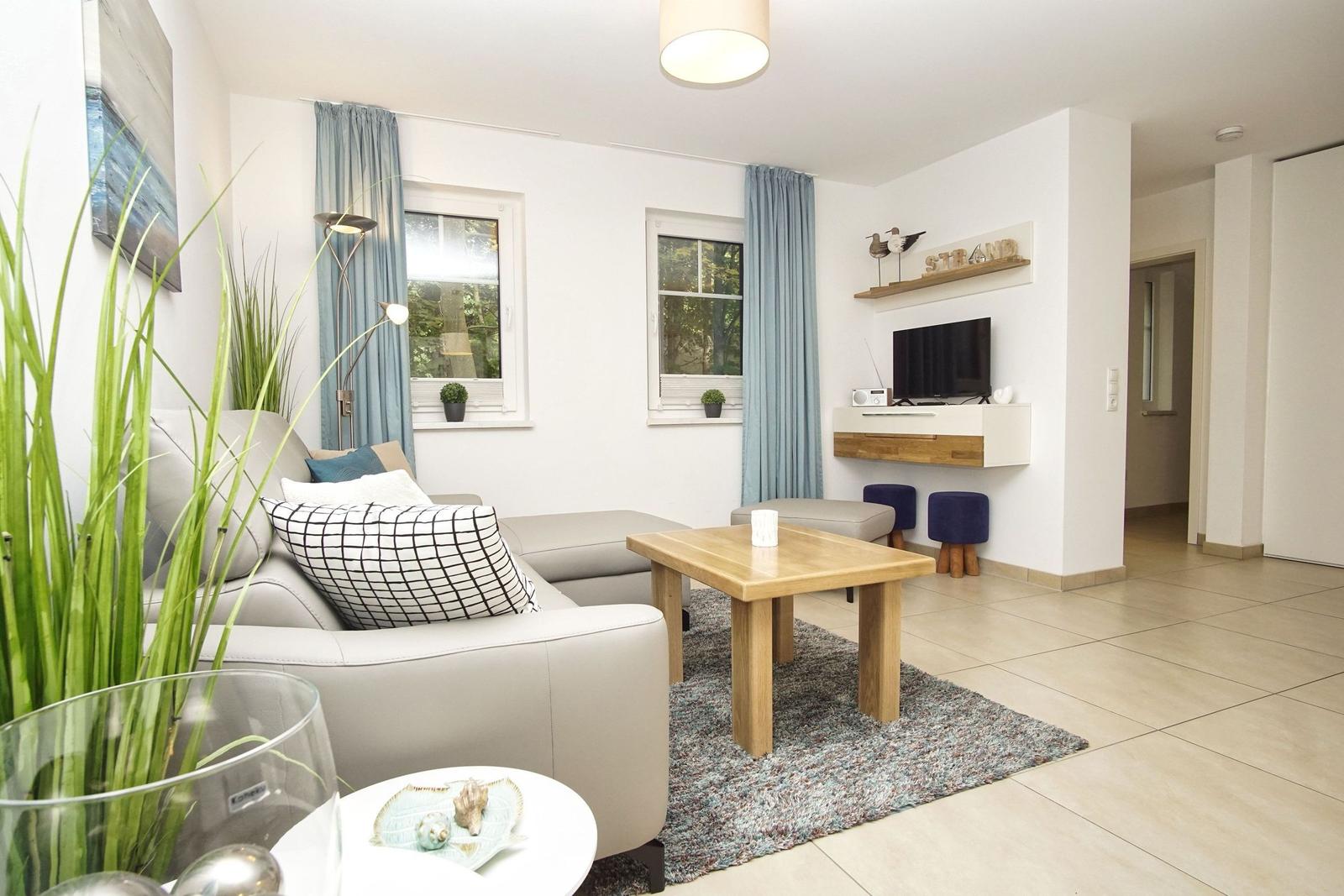 Wohnzimmer mit Sofa, Tisch und Fernseher an Wand. Fenster mit blauen Vorhängen.