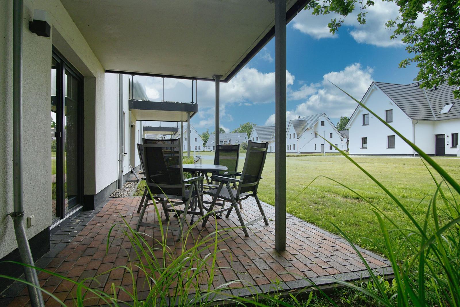 Terrasse mit Tisch und Stühlen, Blick auf grünes Gras und weiße Häuser im Hintergrund.