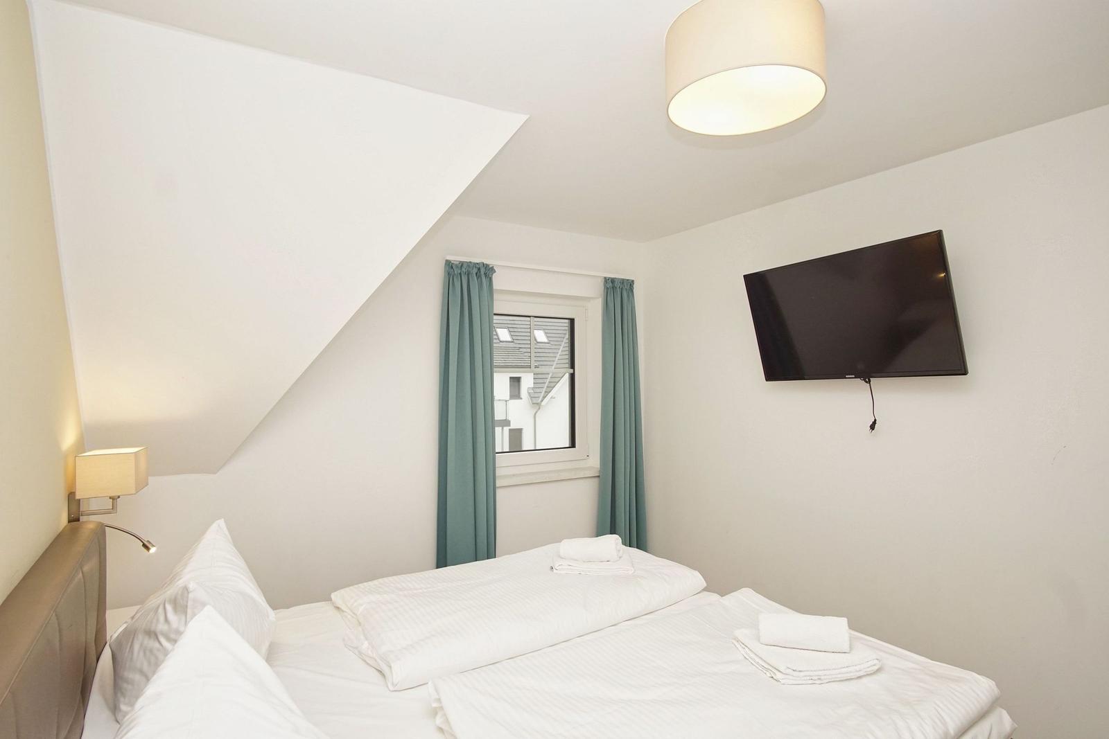 Schlafzimmer mit Doppelbett, Fenster, TV und Deckenleuchte.