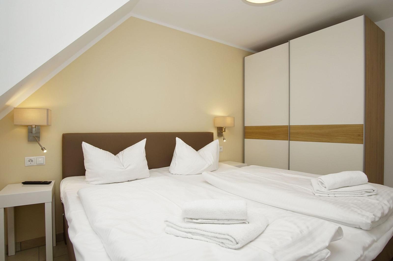 Double bed with white linens and towels, nightstands and wardrobe under sloped ceiling.