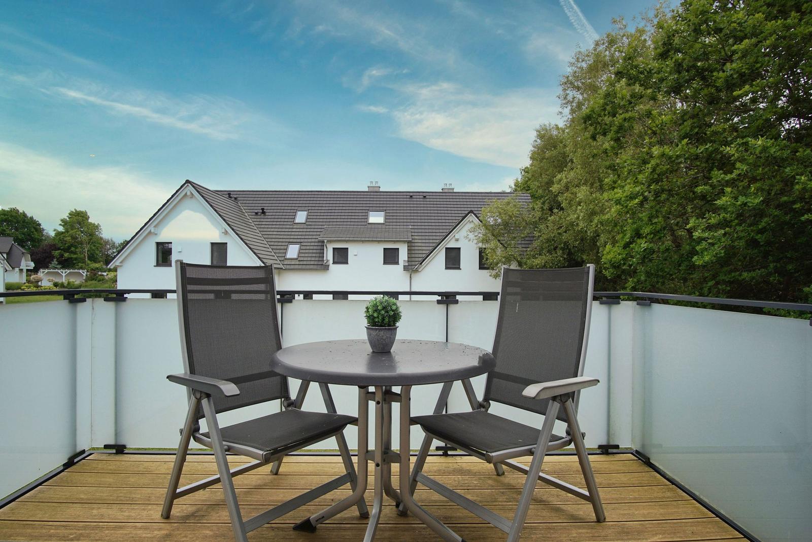 Terrace with table and two chairs. Background: white house and trees.