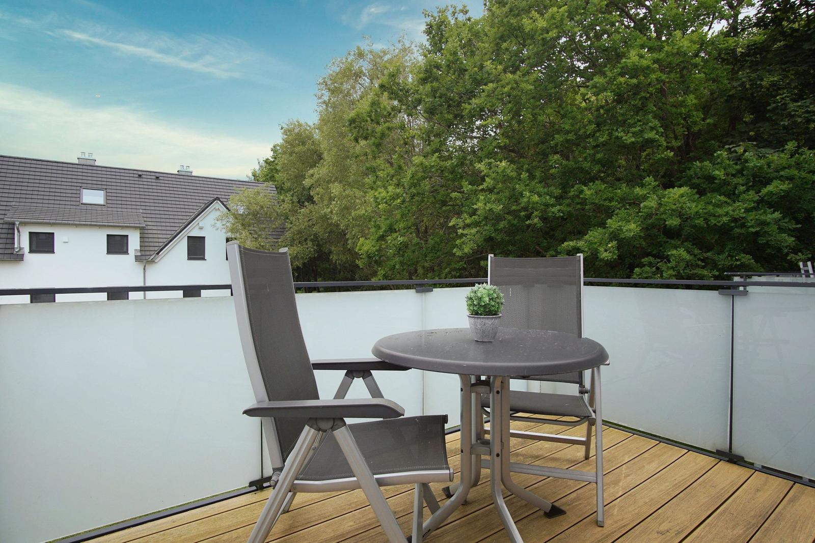 Terrace with table and two chairs, surrounded by trees and a house in the background.